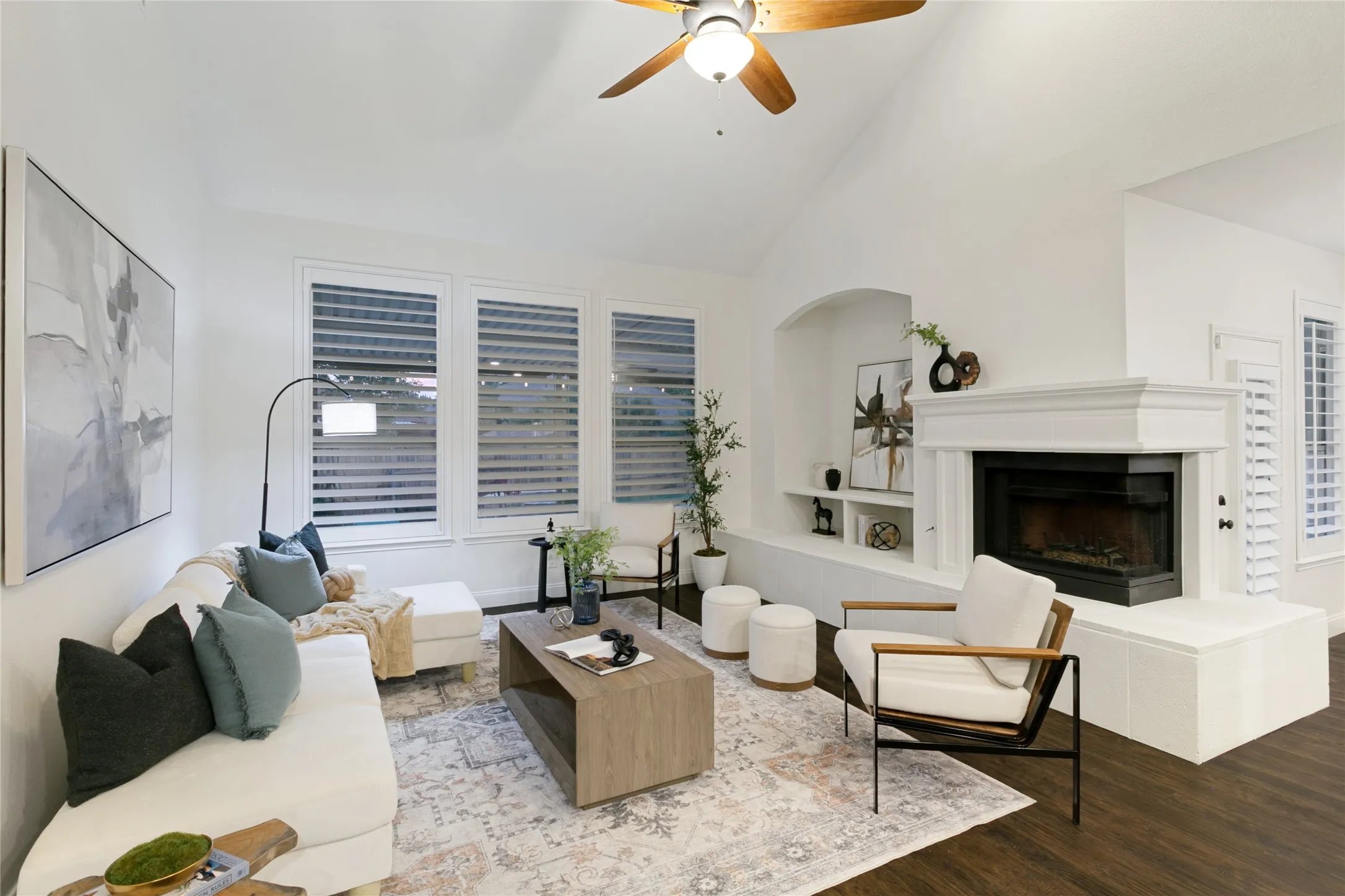 Vaulted ceiling with a ceiling fan, fireplace with a decorative mantel, built-in shelving, wood-finish flooring, and window shutters