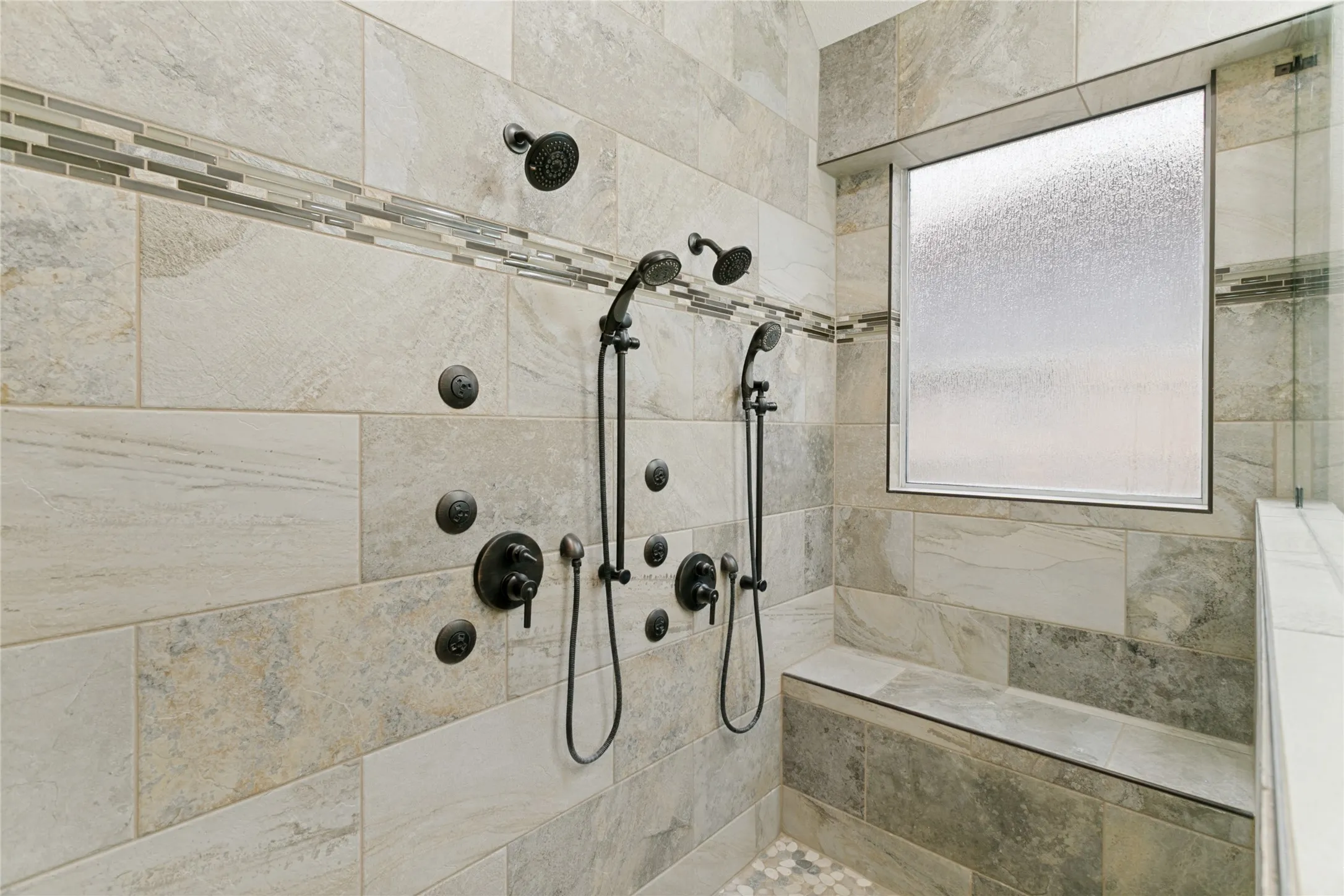 Spacious shower with dual shower heads, body sprayers, integrated bench seating, and mixed material tilework including pebble flooring