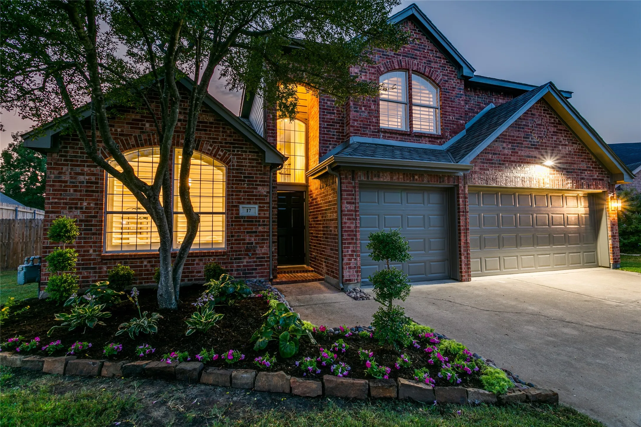 Brick exterior with arched windows, an attached Three-car garage, and a landscaped front yard featuring mature trees and planted flower beds