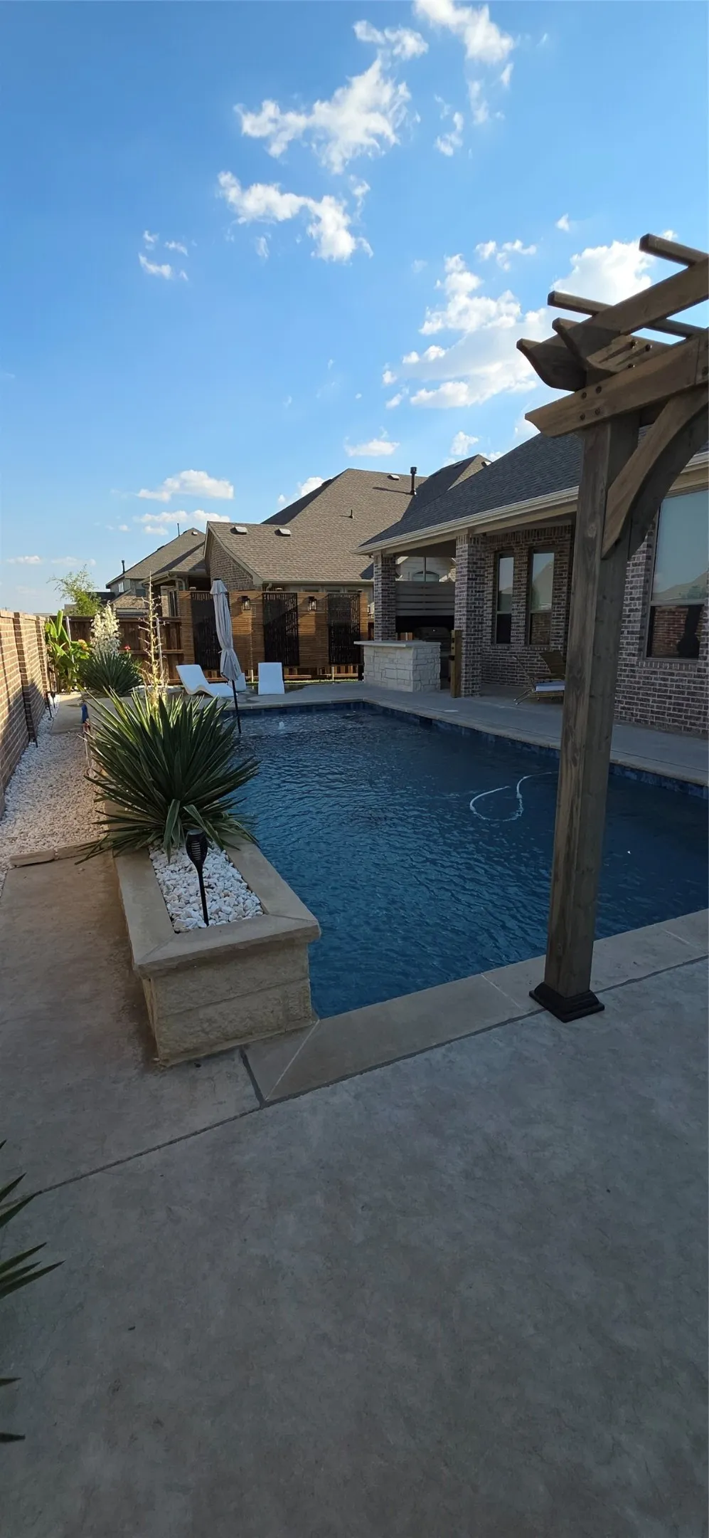 In-ground swimming pool, concrete patio, built-in outdoor bar, stone planter, and wooden pergola