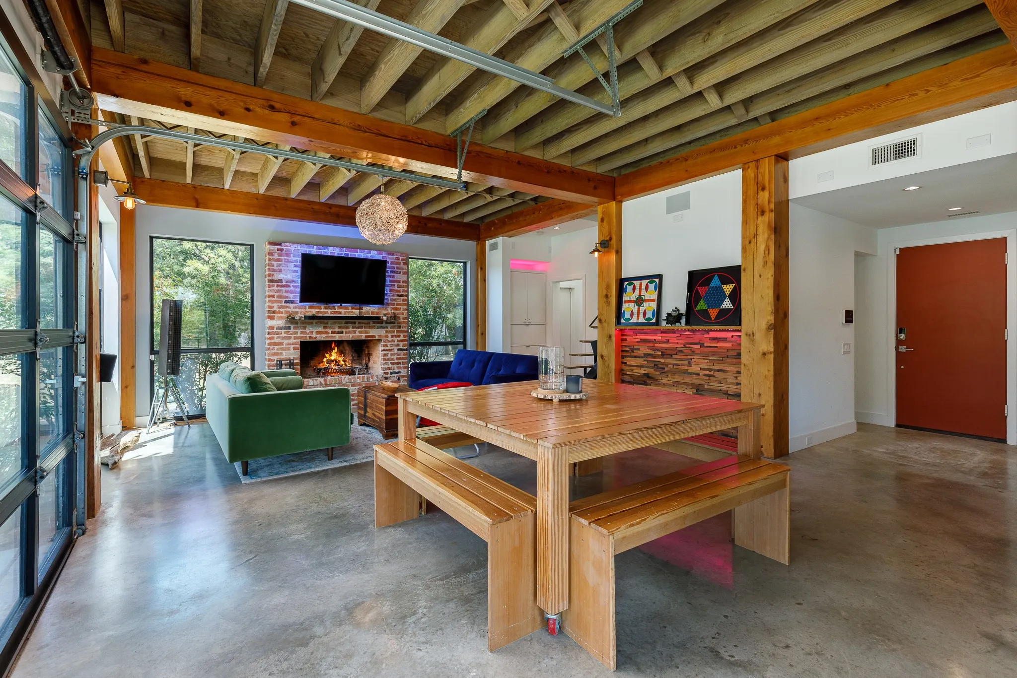 Exposed wood beam ceiling, brick fireplace, concrete flooring, full-light glass garage door, and a built-in wood accent wall