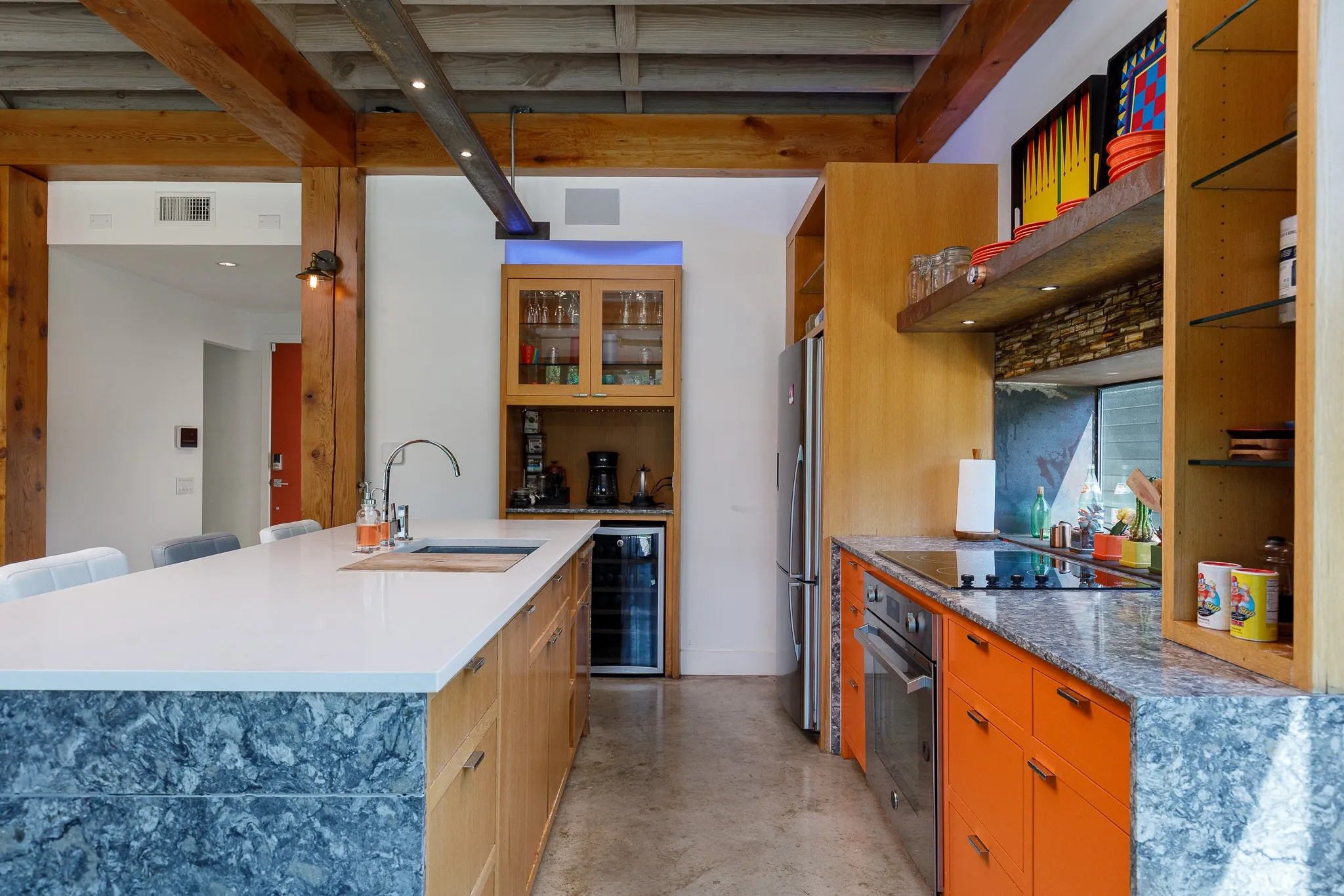 Exposed wood beams, concrete flooring, stainless steel appliances, white countertop island, and vibrant orange cabinetry