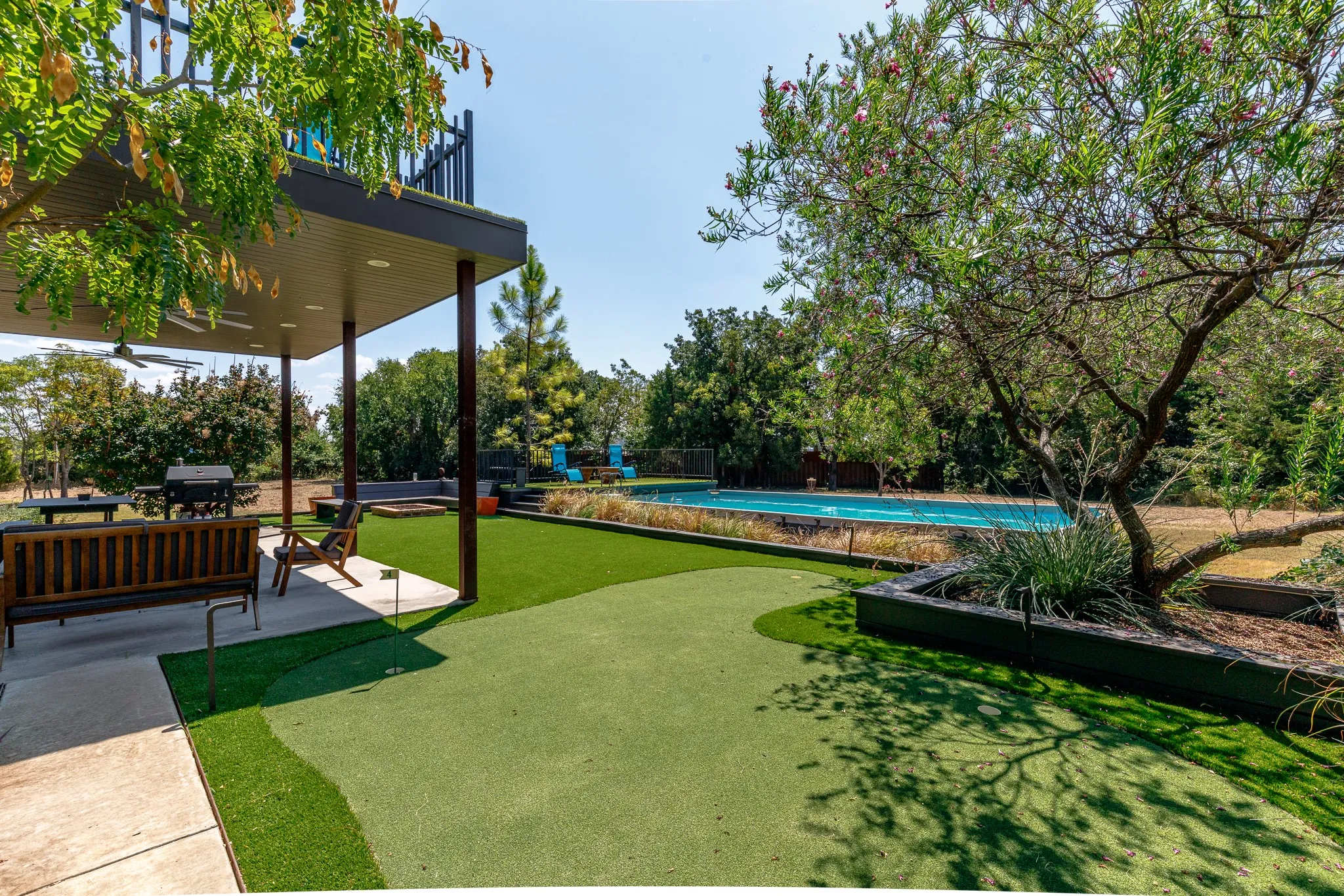 Covered patio with ceiling fan, artificial turf putting green, swimming pool, mature trees, and 1 of 5 puttin greens