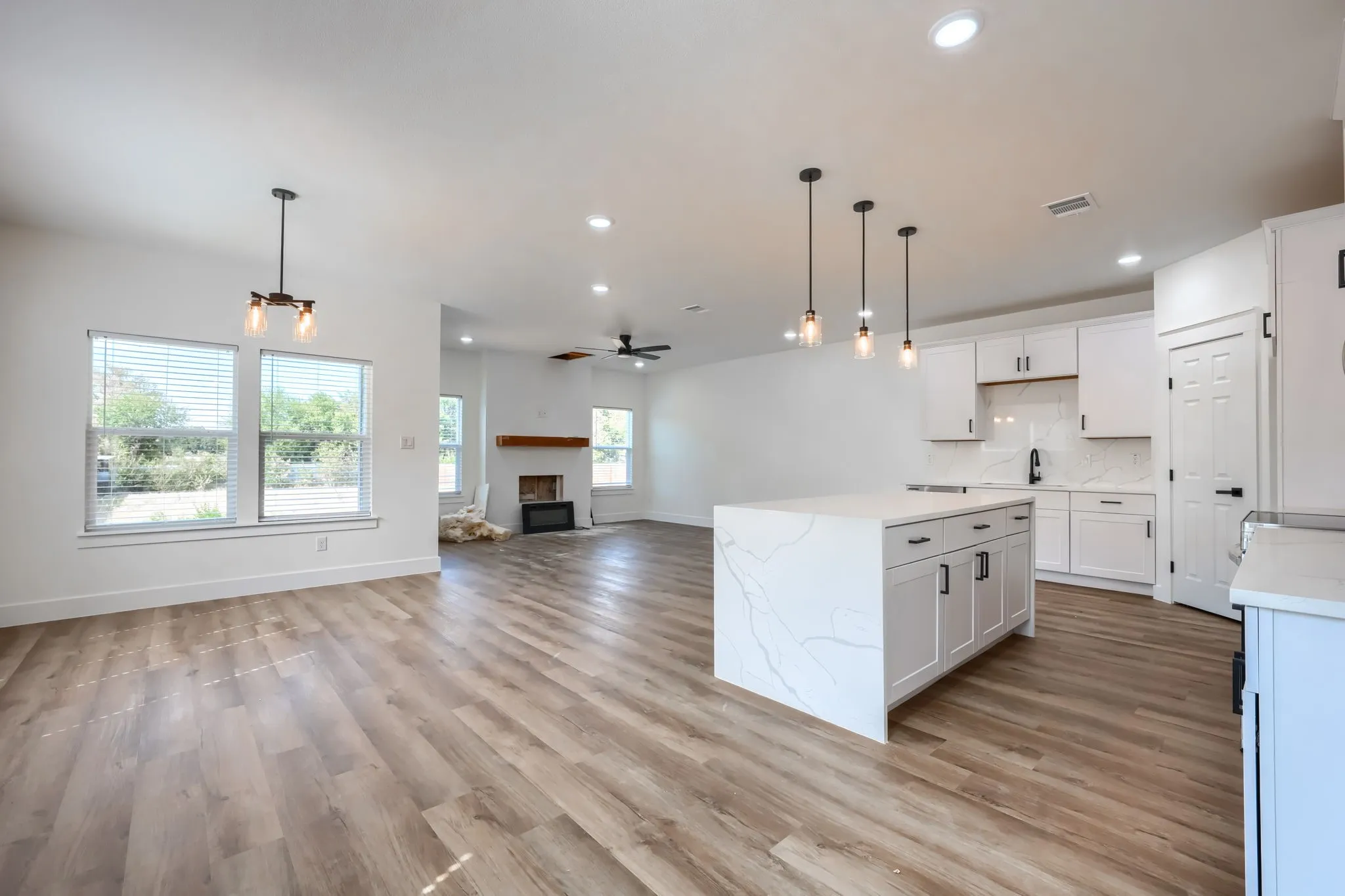 Open concept living area featuring wood-finish flooring, a white kitchen island with a waterfall countertop, white cabinetry with black hardware, and an integrated fireplace with a wood mantle