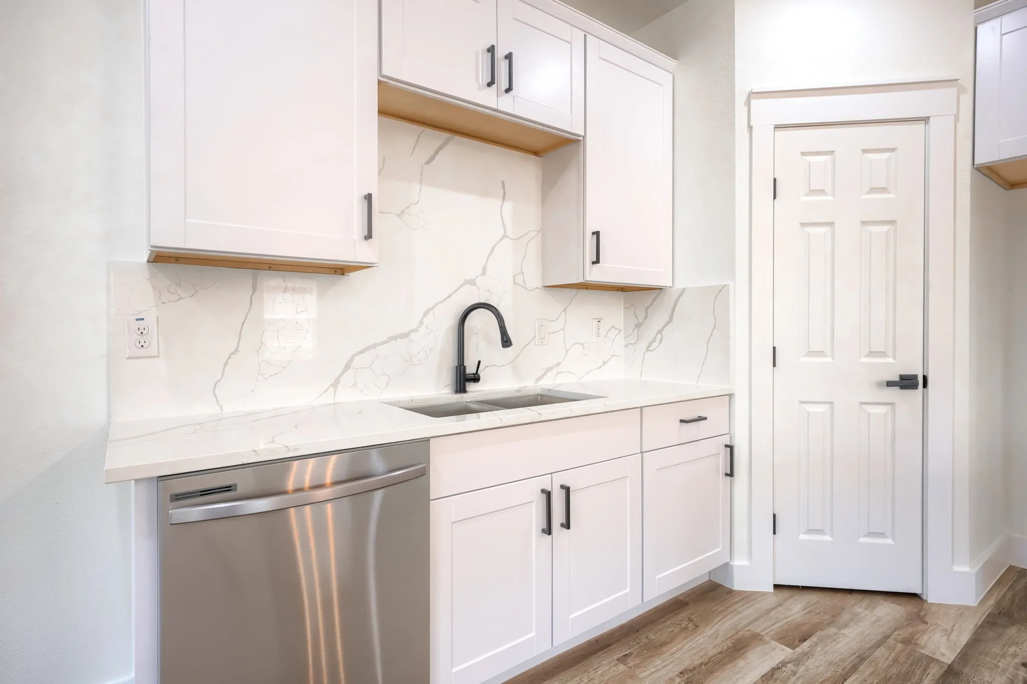 White shaker cabinetry with matte black hardware, integrated stainless steel dishwasher, undermount sink with a matte black faucet, full-height backsplash, and wood-finish flooring