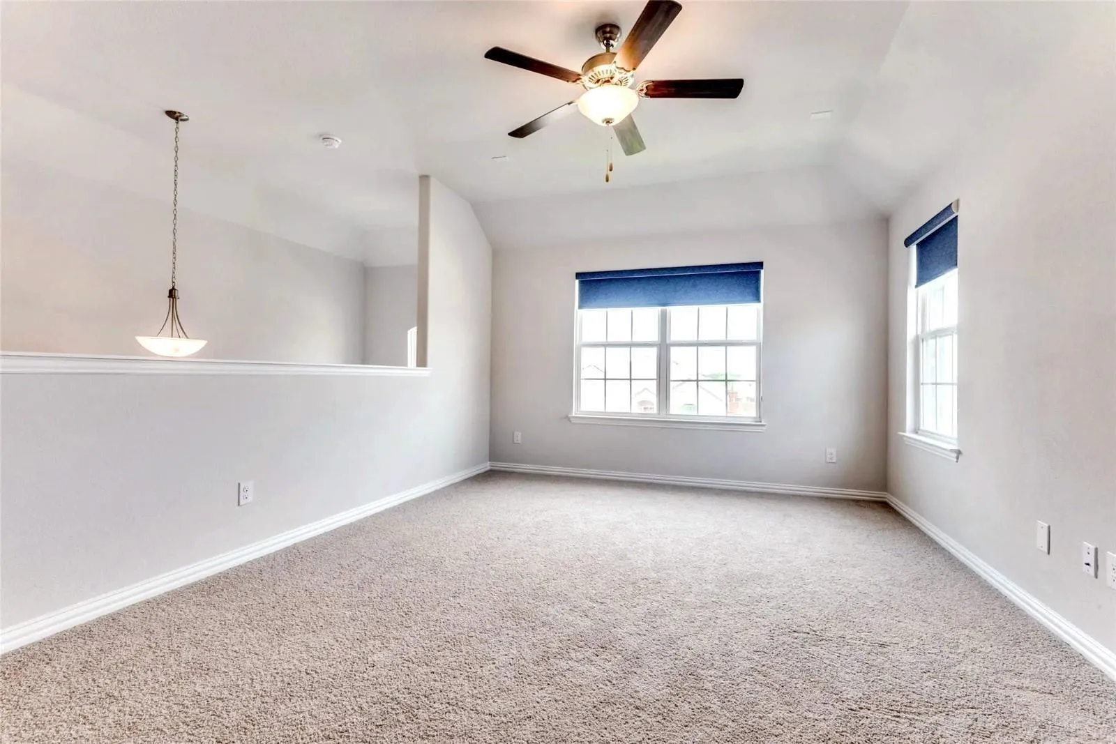 Carpeted interior space featuring a vaulted ceiling with a ceiling fan, multiple windows with roller blinds, and a built-in half-wall partition with an overhead pendant light