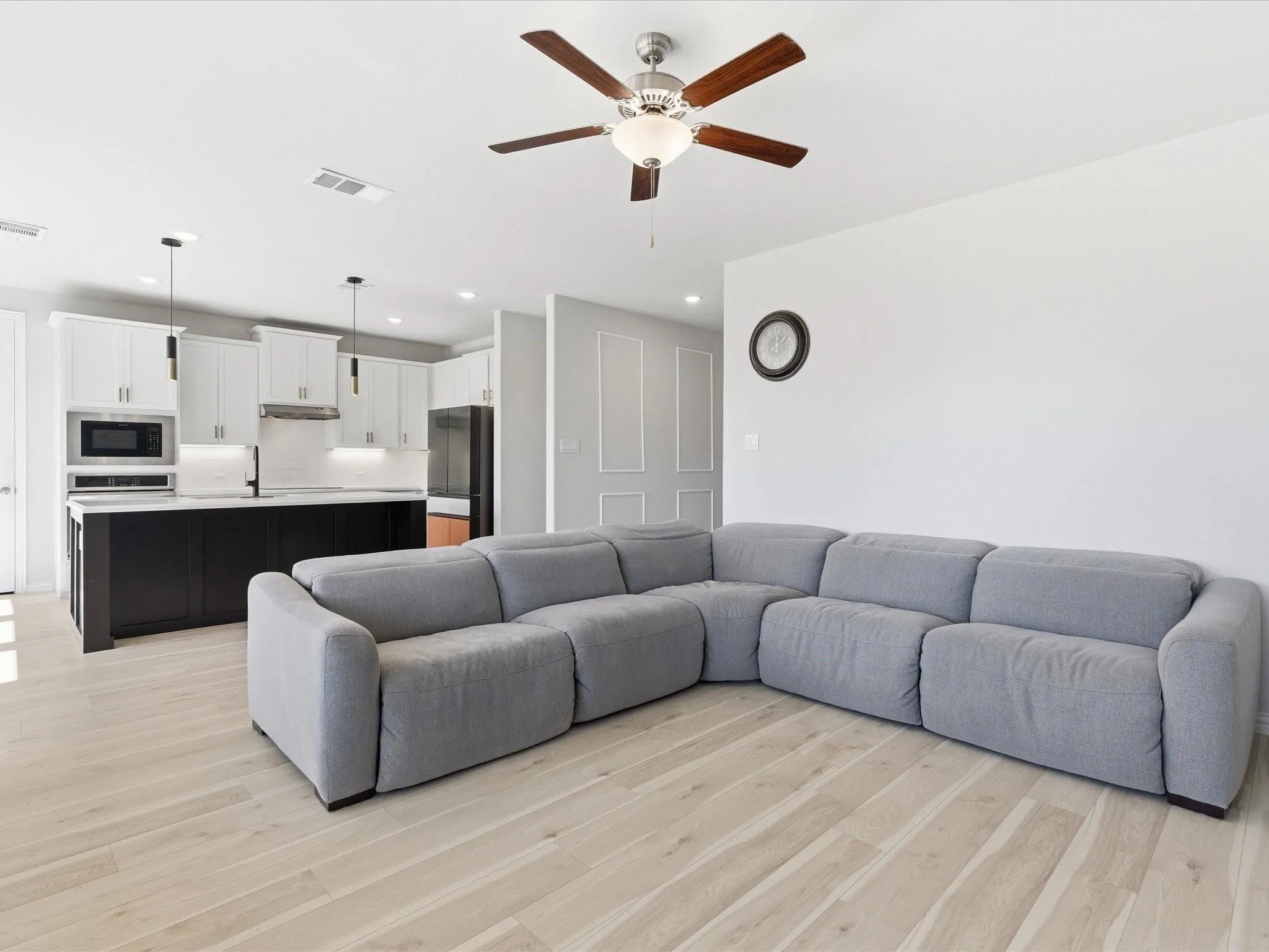 Open-concept living area featuring wood-finish flooring, a ceiling fan with light, and a kitchen with white upper cabinetry, a dark island, and pendant lighting