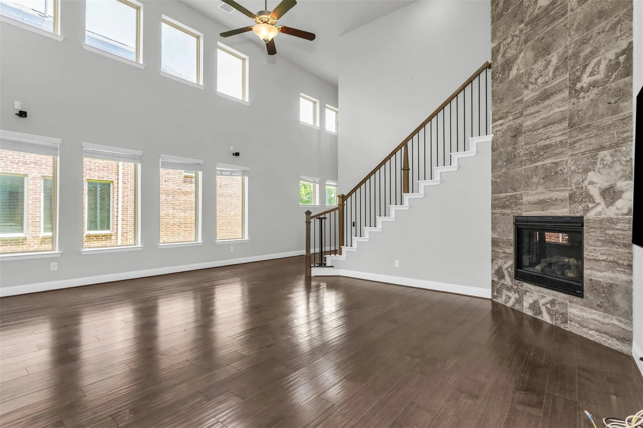 Two-story ceiling, wood-finish flooring, staircase with metal balusters, tiled fireplace with glass enclosure, and ceiling fan with integrated lighting