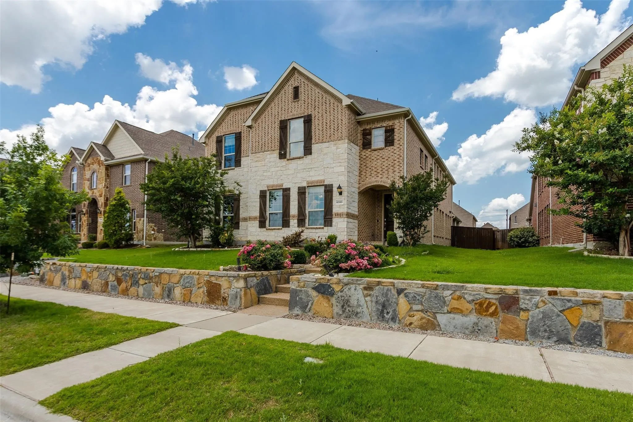 Two-story residence featuring a brick and stone facade, dark window shutters, a stone retaining wall, and landscaped front yard with mature trees