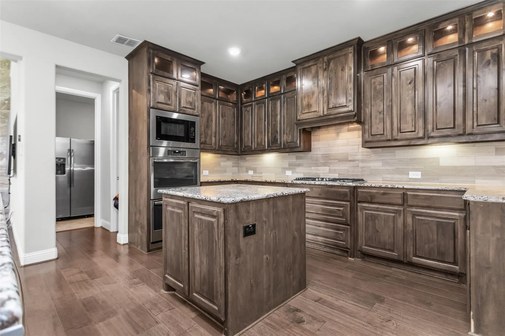 Kitchen featuring extensive wood cabinetry, wood-finish flooring, a central island, stainless steel built-in appliances, and light-toned tile backsplash