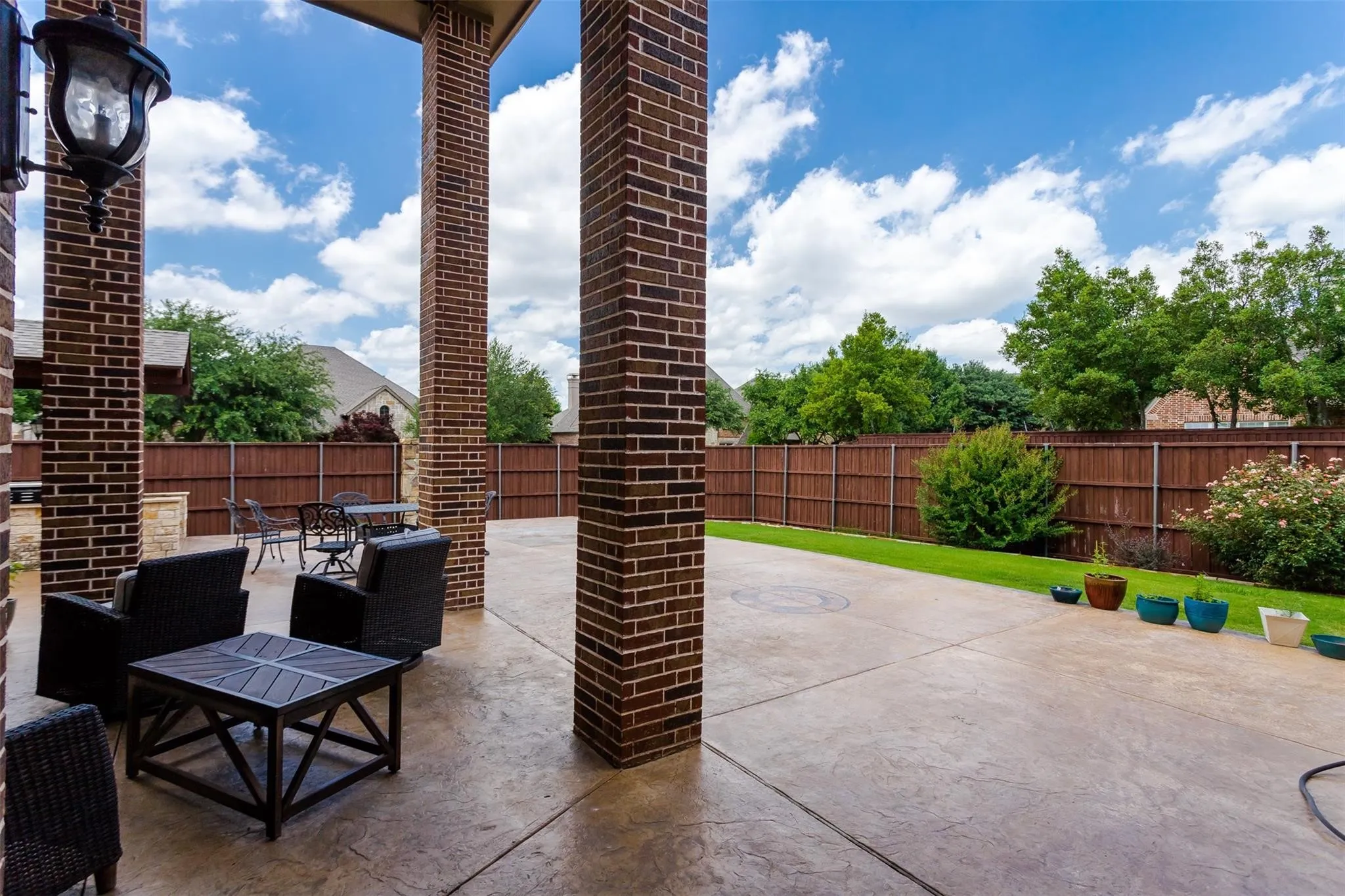 Brick columns supporting a covered patio with stamped concrete flooring, leading to a fenced yard with a manicured lawn and mature landscaping