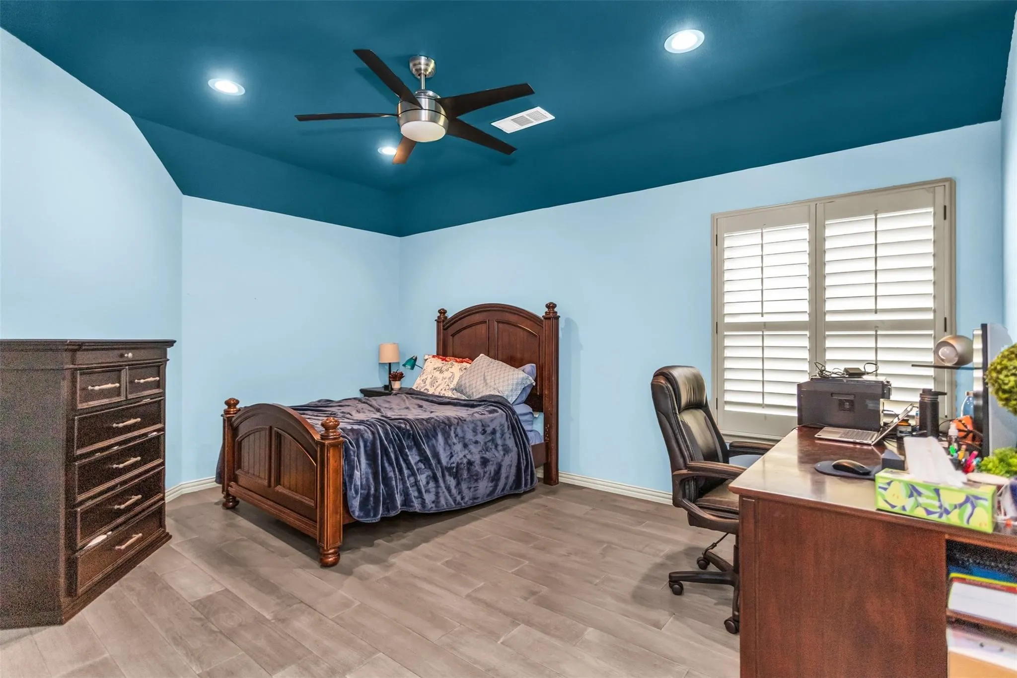 Wood-finish flooring with recessed lighting, a ceiling fan, and a window featuring plantation shutters