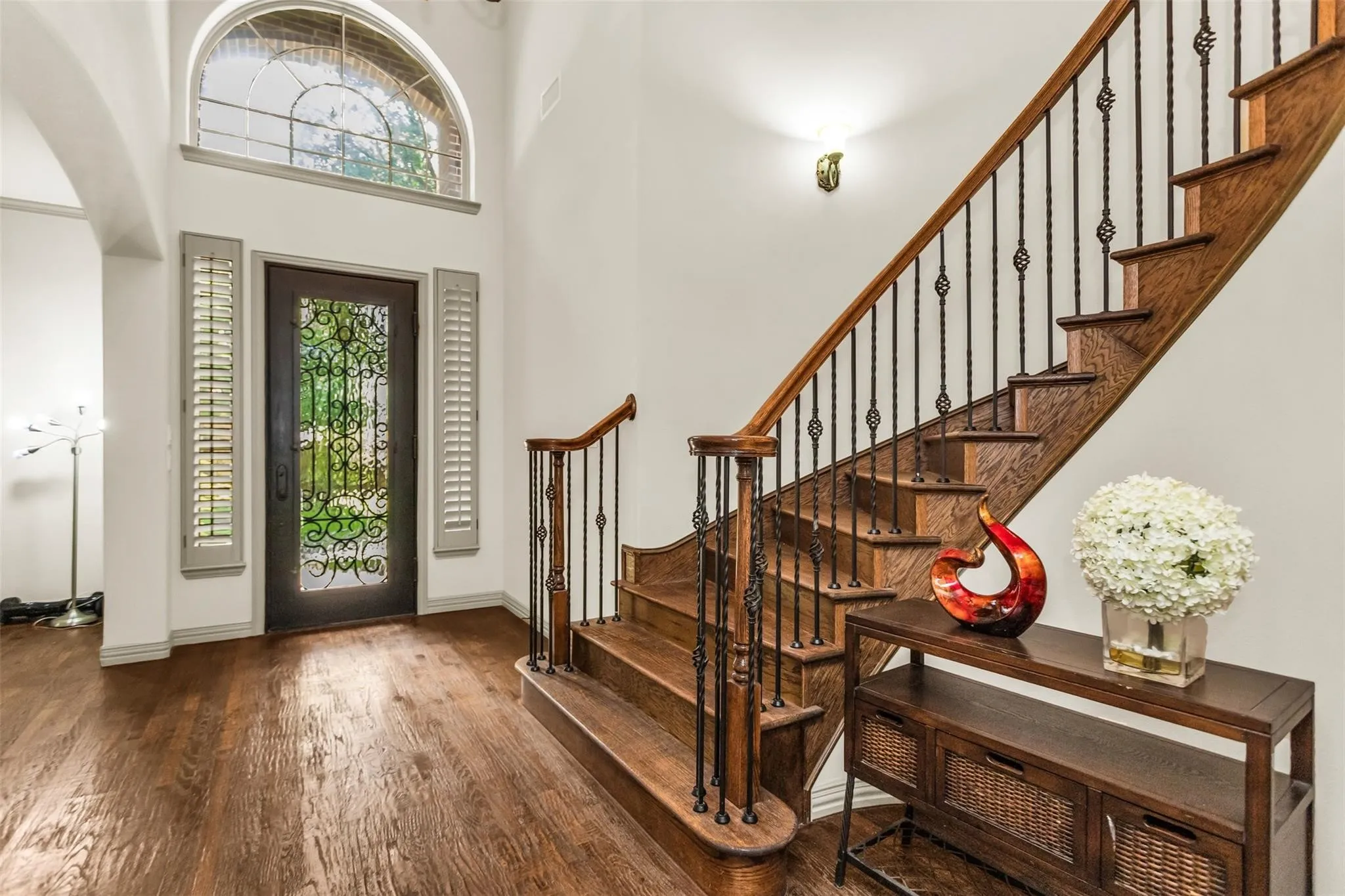 Wood-finish flooring throughout, grand entry door with decorative ironwork, arched transom window, built-in plantation shutters, and a wood staircase with wrought iron balusters