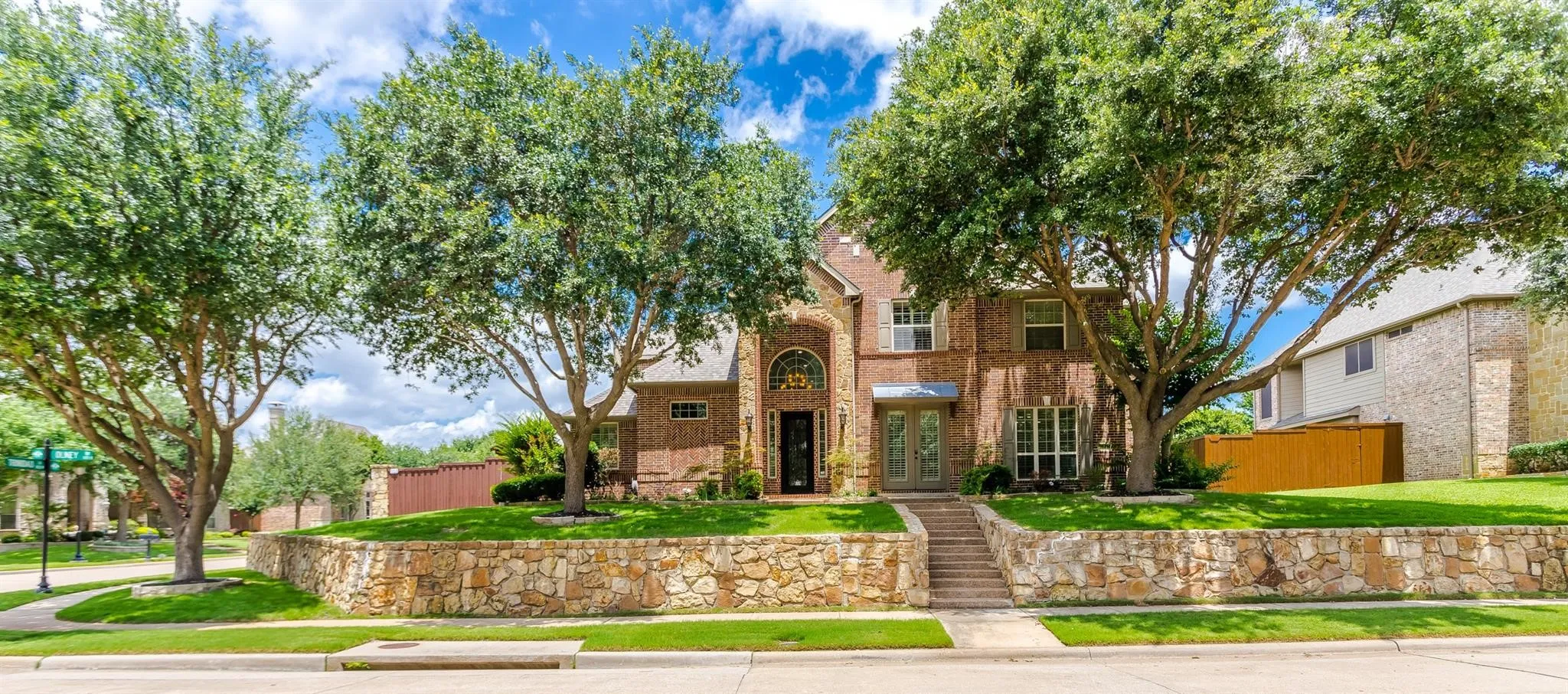 Brick exterior with arched entryway, stone retaining wall, mature trees, and a concrete walkway with stone steps