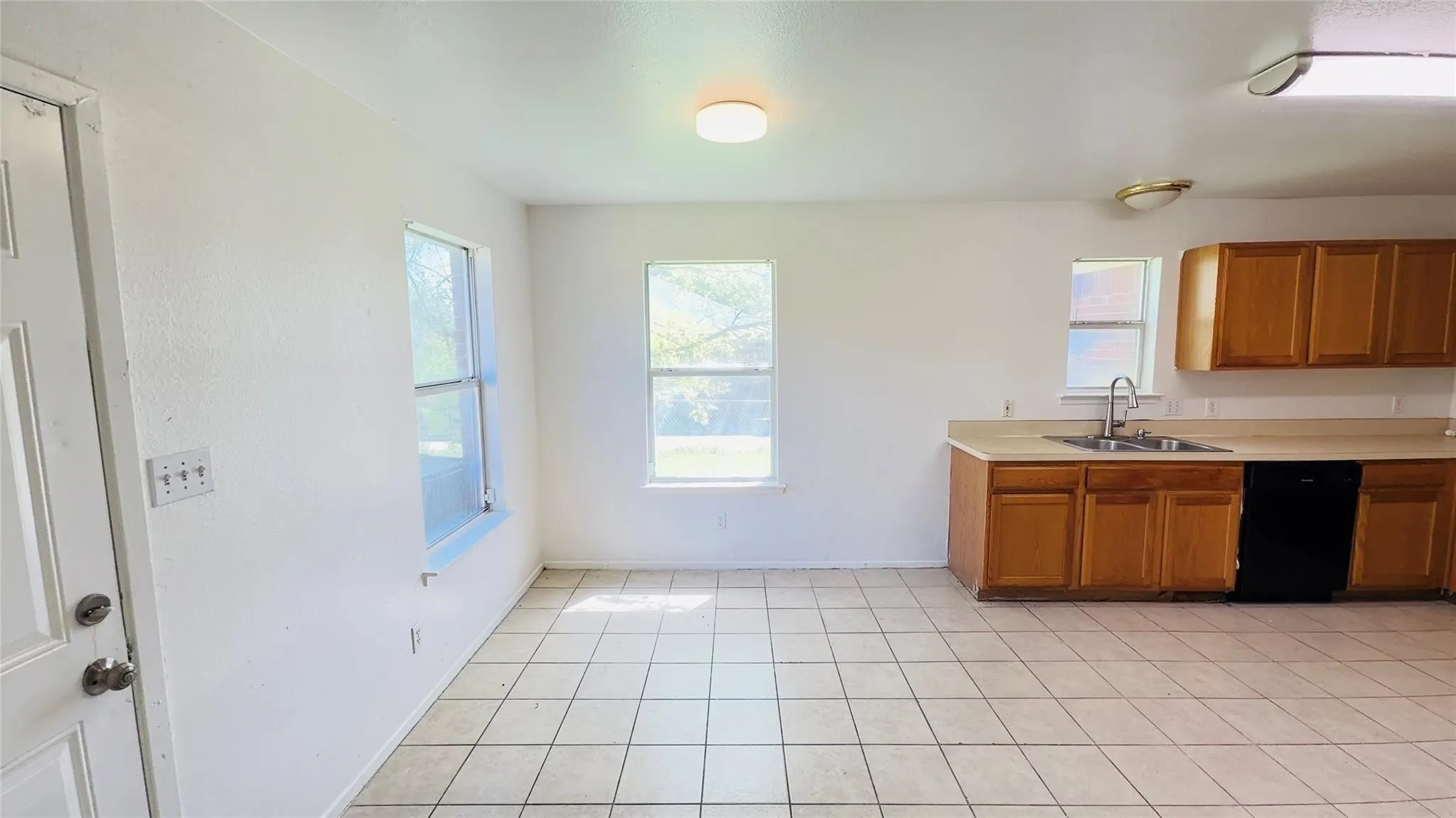 Spacious open-concept area featuring tile flooring, multiple windows, kitchen cabinetry with a built-in sink, and a black dishwasher