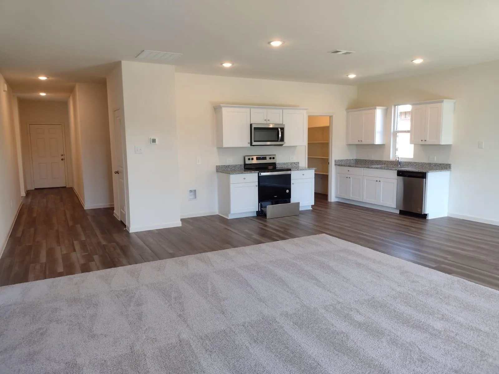Open-concept living space featuring wood-finish flooring, recessed lighting, and a kitchen with white cabinetry, stainless steel appliances, and a pantry with shelving