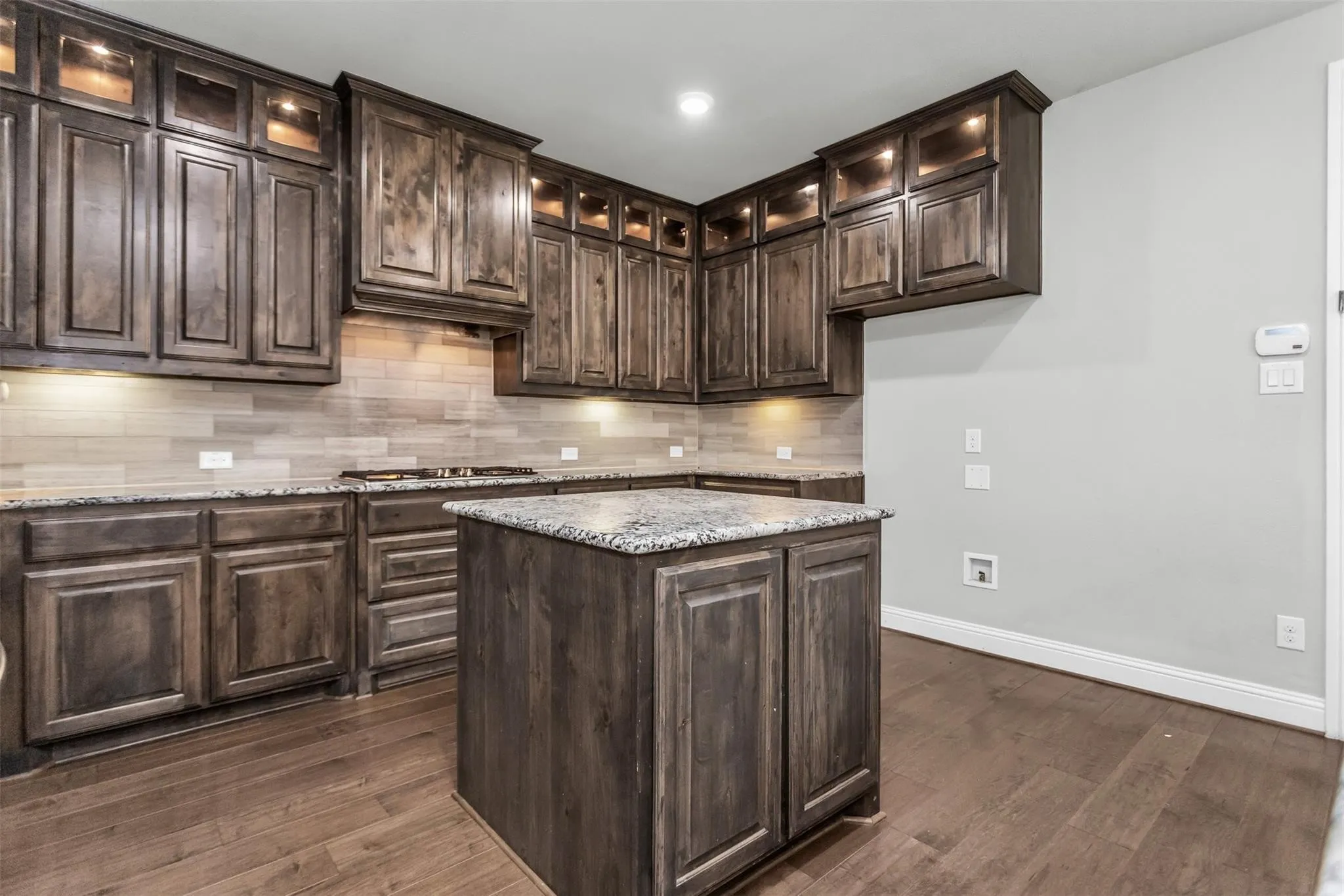 Wood-finish flooring, dark wood cabinetry with integrated lighting, a central island with a stone countertop, a tile backsplash, and built-in appliances