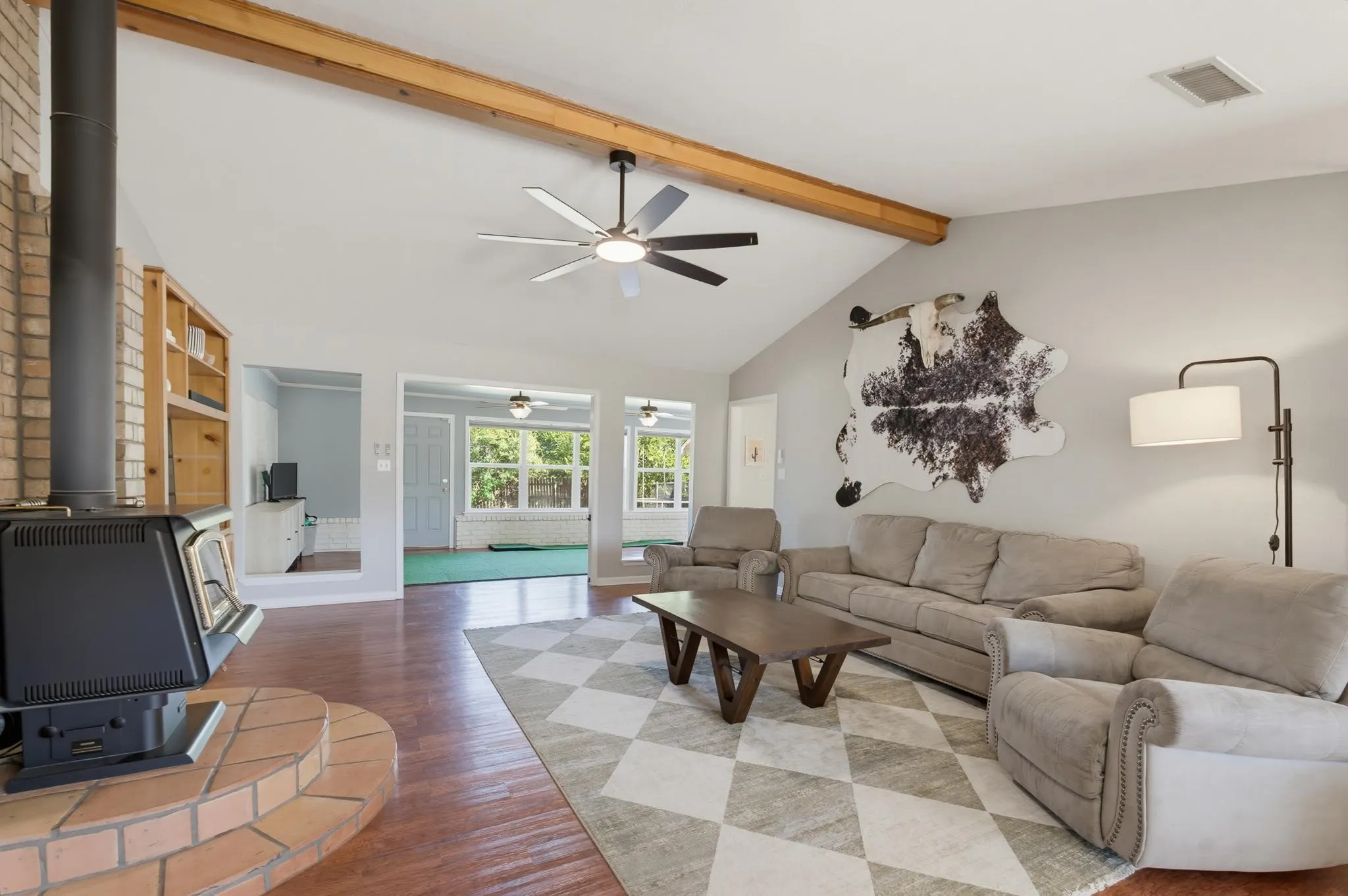 Vaulted ceiling with exposed beam, ceiling fan, wood-finish flooring, brick-surround wood-burning stove, and built-in shelving