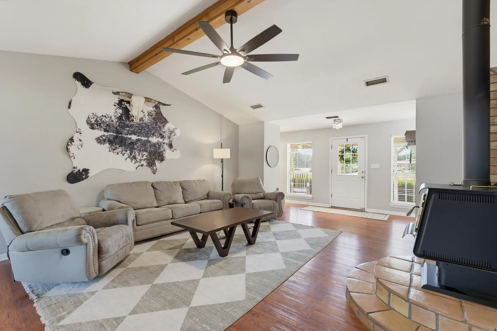 Vaulted ceiling with exposed wood beams, wood-finish flooring, ceiling fan with integrated lighting, exposed brick and metal fireplace, and a white panel door