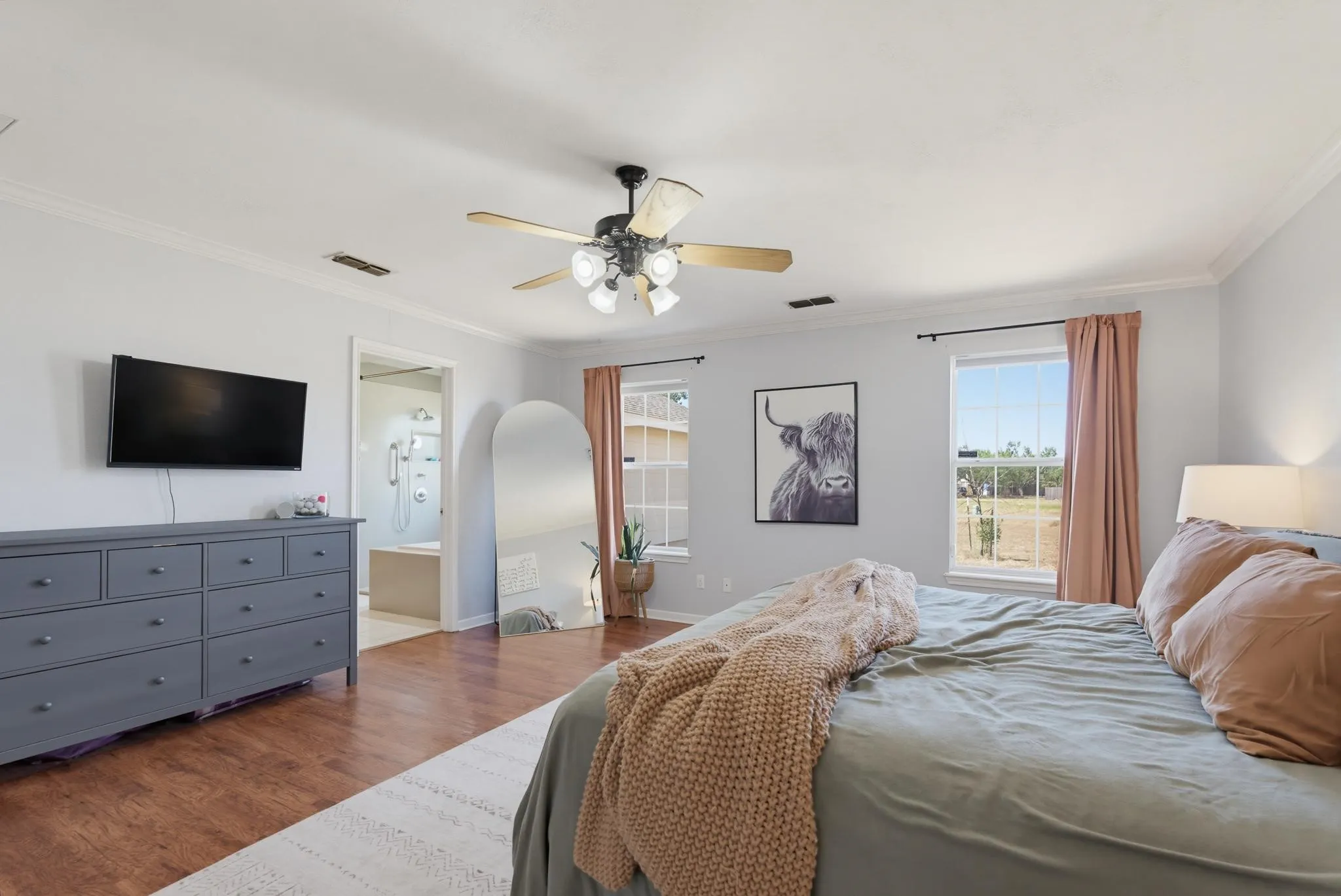 Spacious primary bedroom with wood-finish flooring, ceiling fan with integrated lighting, dual dressers with ample storage, and multiple windows providing natural light