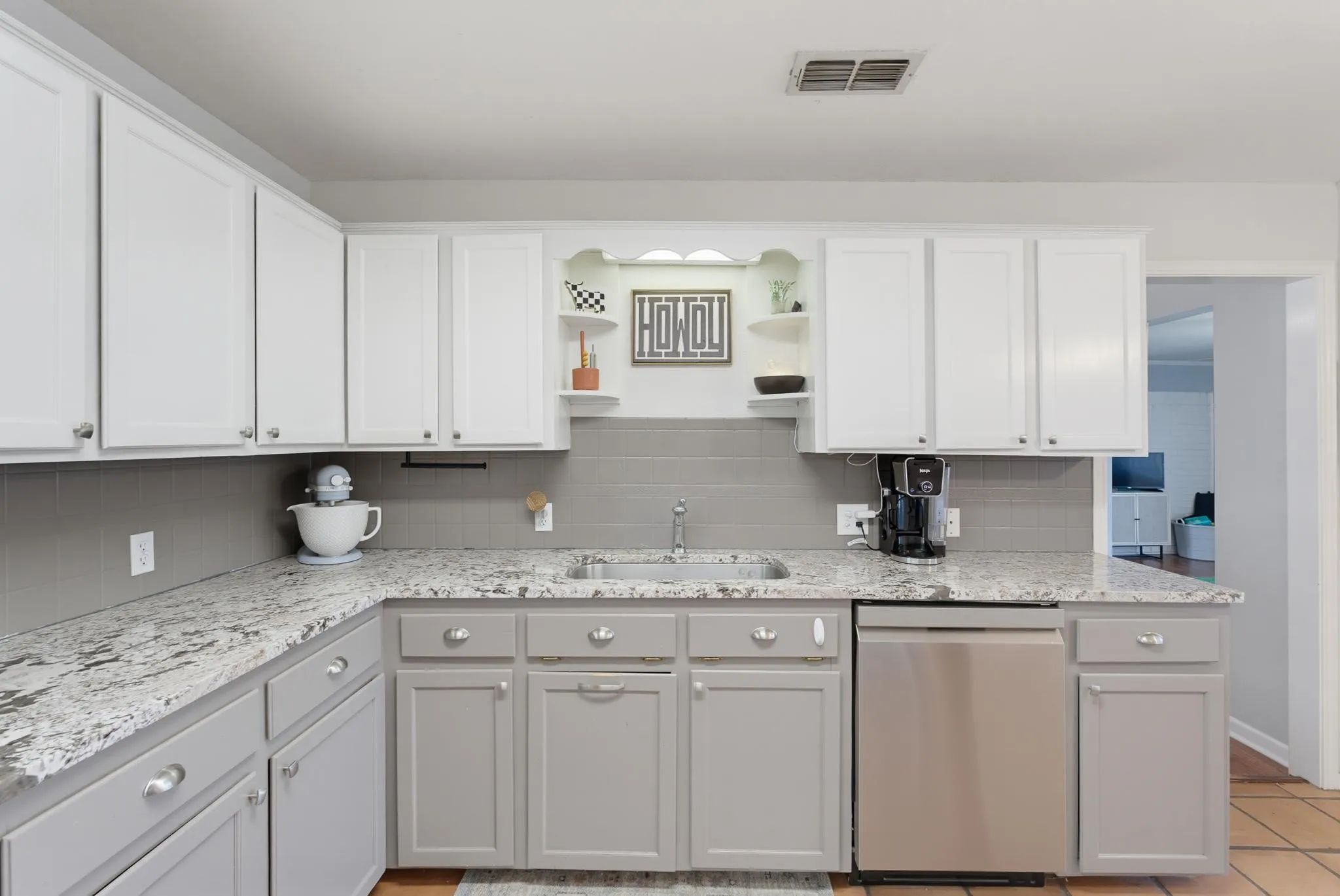 White upper cabinetry, gray lower cabinetry, light-toned countertops, gray tile backsplash, and a stainless steel dishwasher