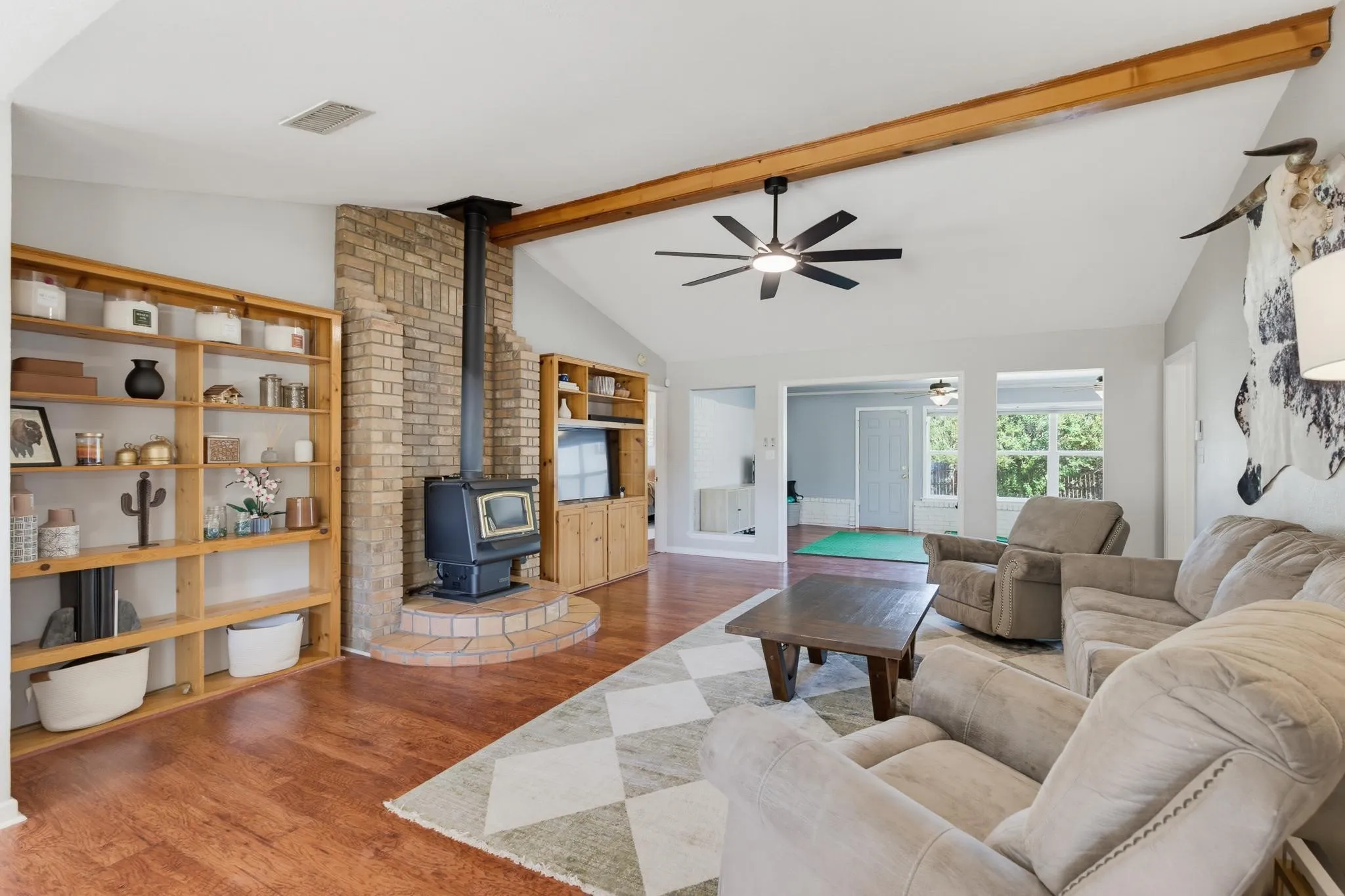 Vaulted ceiling with an exposed wood beam, wood-finish flooring, brick-clad wood-burning stove, built-in shelving, and a ceiling fan with integrated lighting