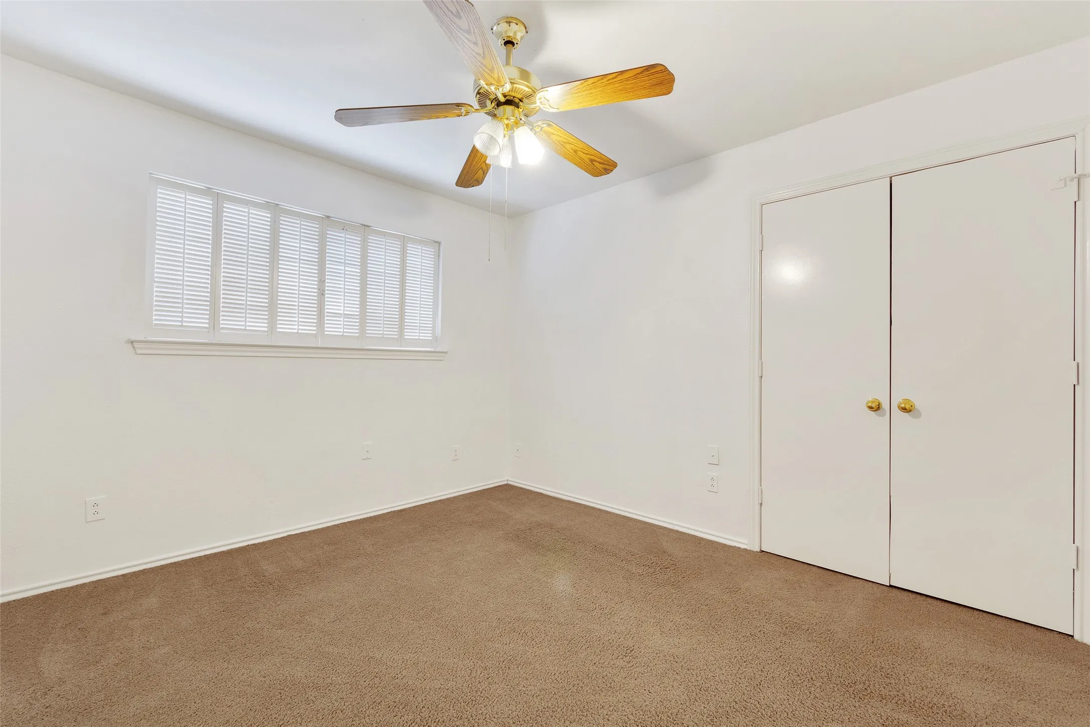Recessed window with white blinds, neutral wall paint, ceiling fan with light fixture, double-door closet with gold-finish hardware, and carpet flooring