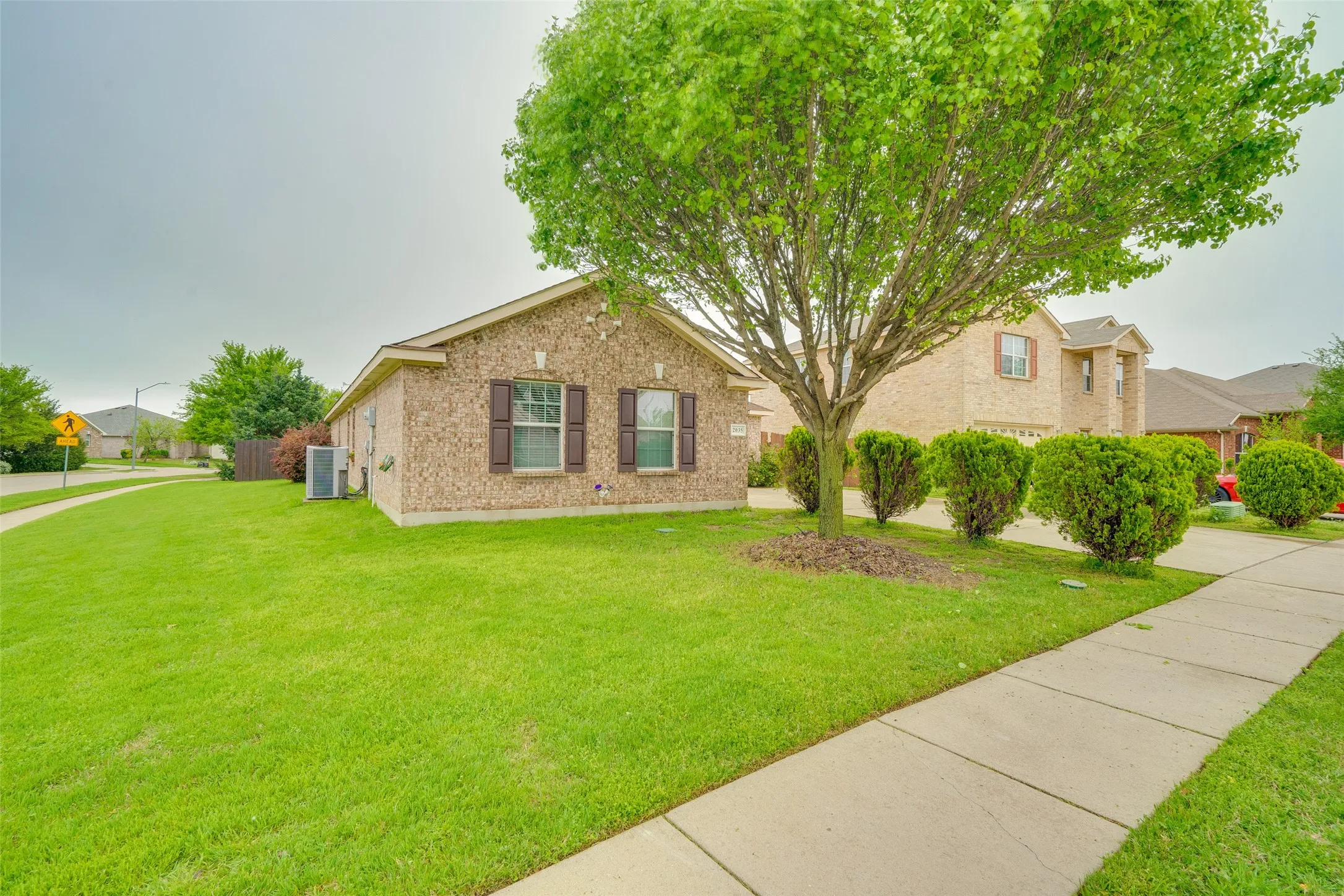 Brick exterior with brown shutters, mature shade tree, manicured shrubs, and a concrete sidewalk
