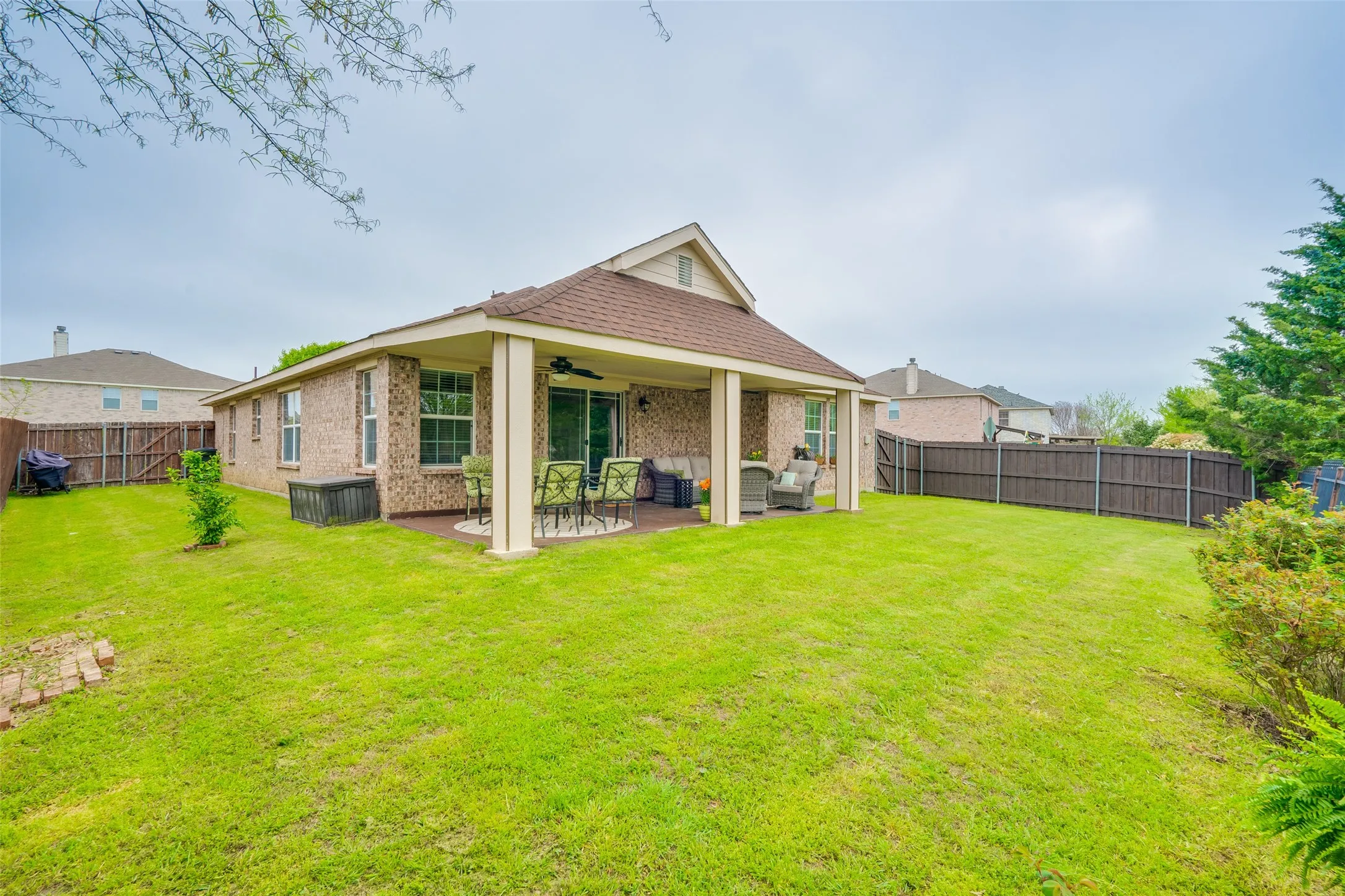 Expansive green lawn, covered patio with column supports, brick exterior, and full privacy fencing