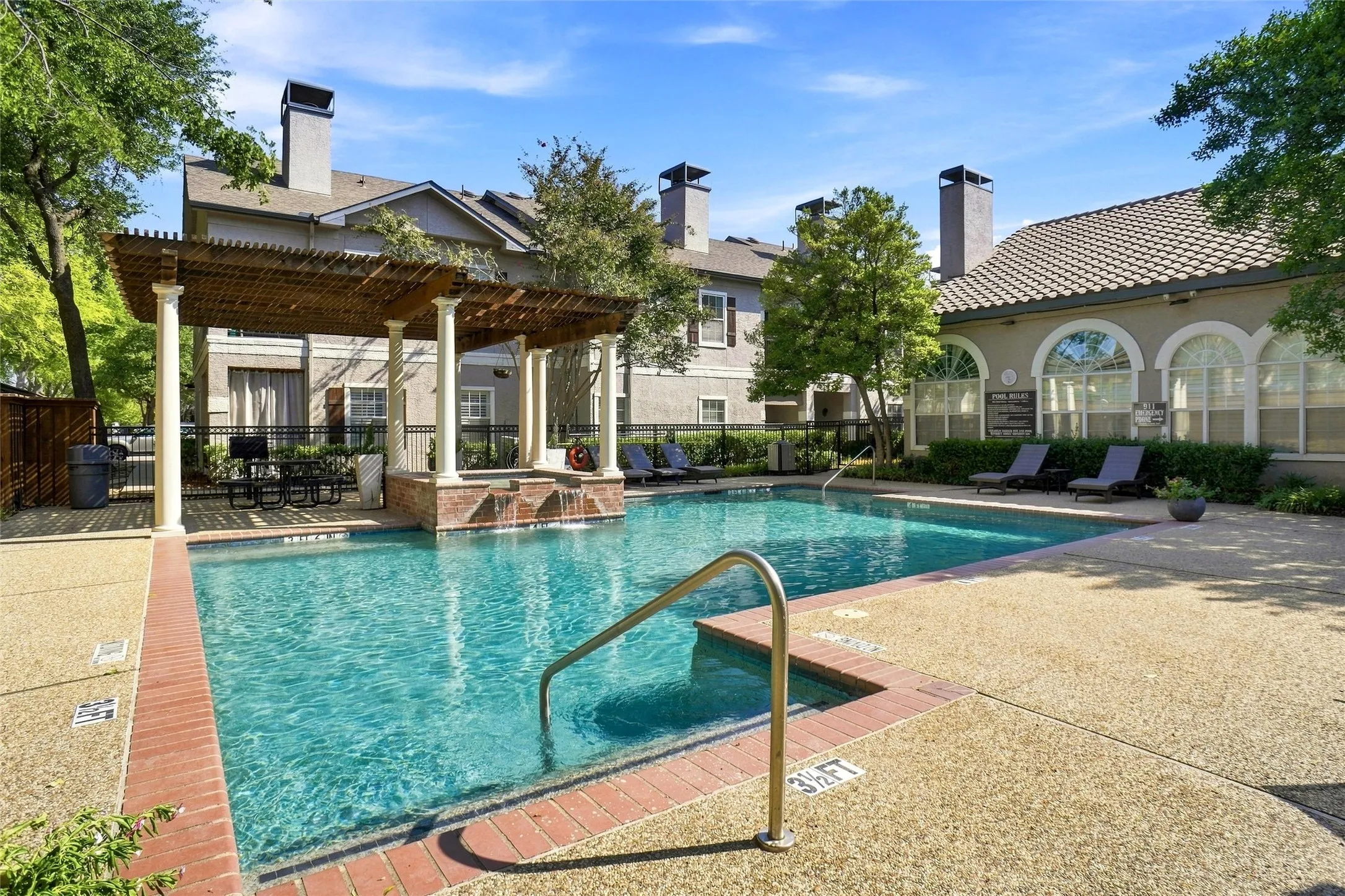 Outdoor swimming pool with integrated spa and water feature, surrounded by exposed aggregate concrete decking and brick coping