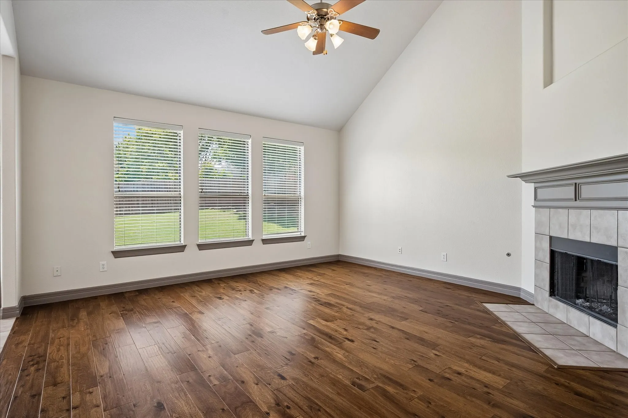 Wood-finish flooring throughout, vaulted ceiling, ceiling fan with integrated lighting, three windows with blinds, and a tiled fireplace with a mantel