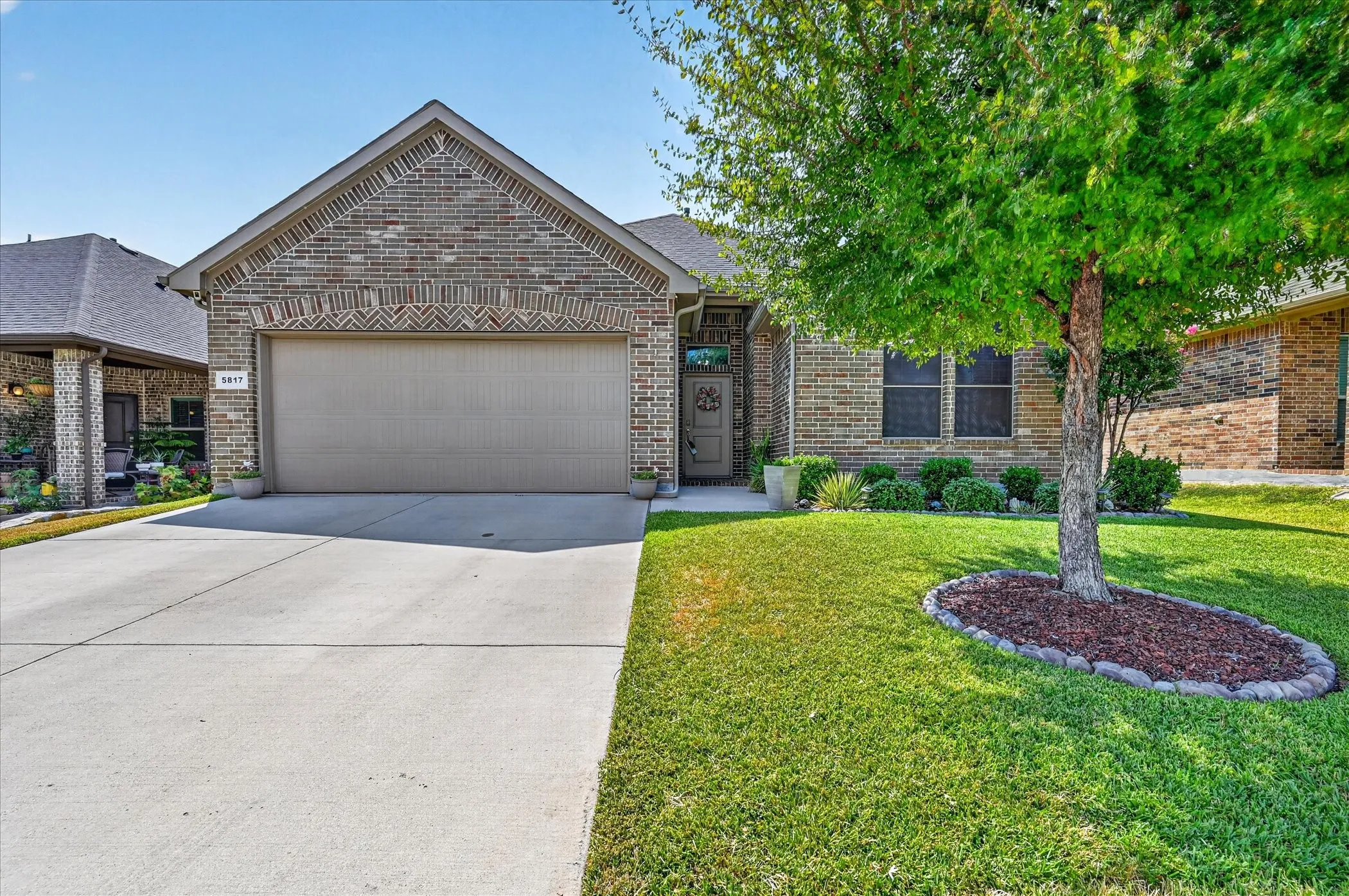 Brick exterior home featuring a two-car garage with a concrete driveway, manicured front lawn, and mature landscaping including a large shade tree with mulched planting bed