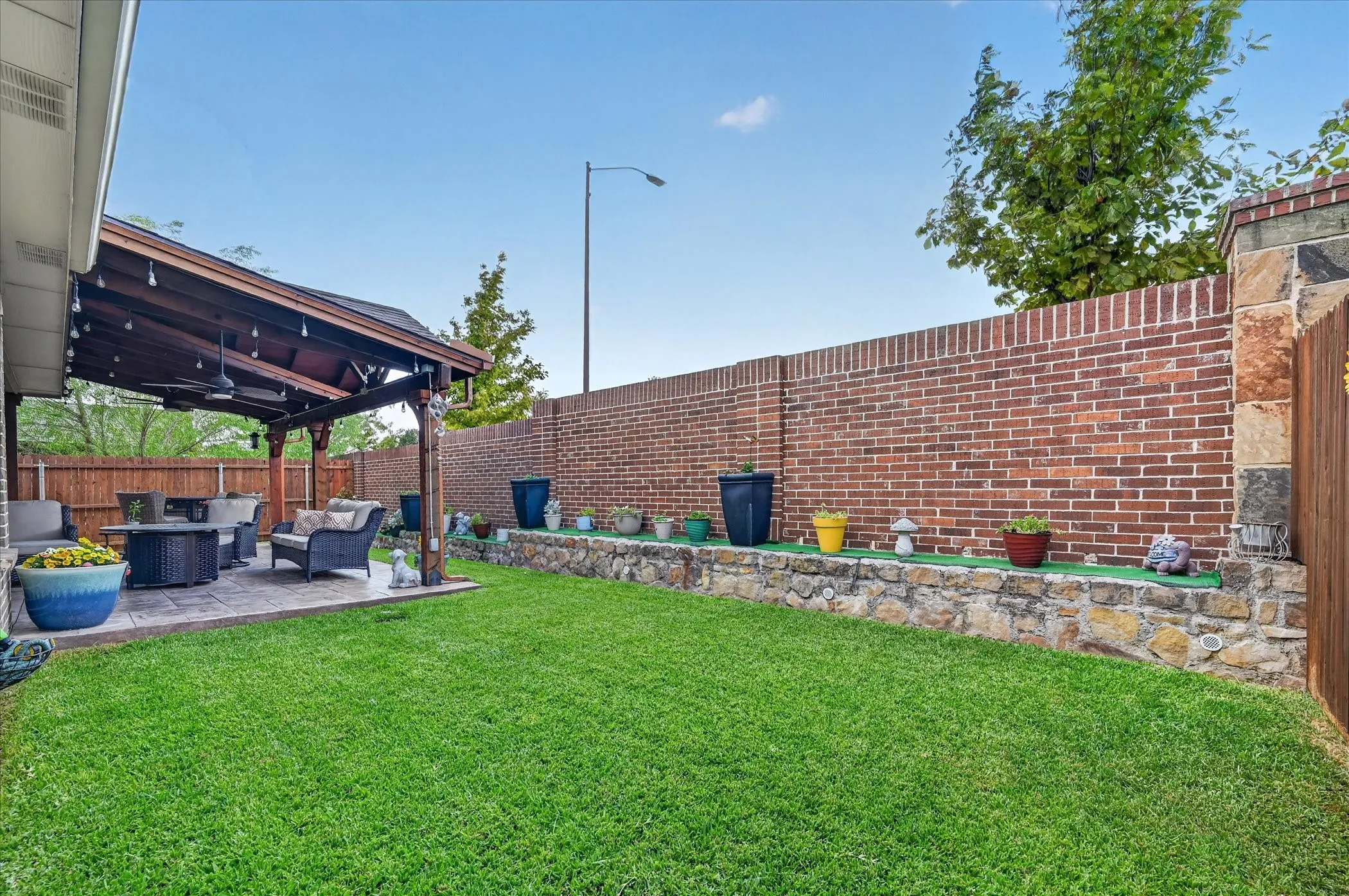 Covered patio with stamped concrete, expansive lawn, stone retaining wall, brick privacy wall, and wood fence