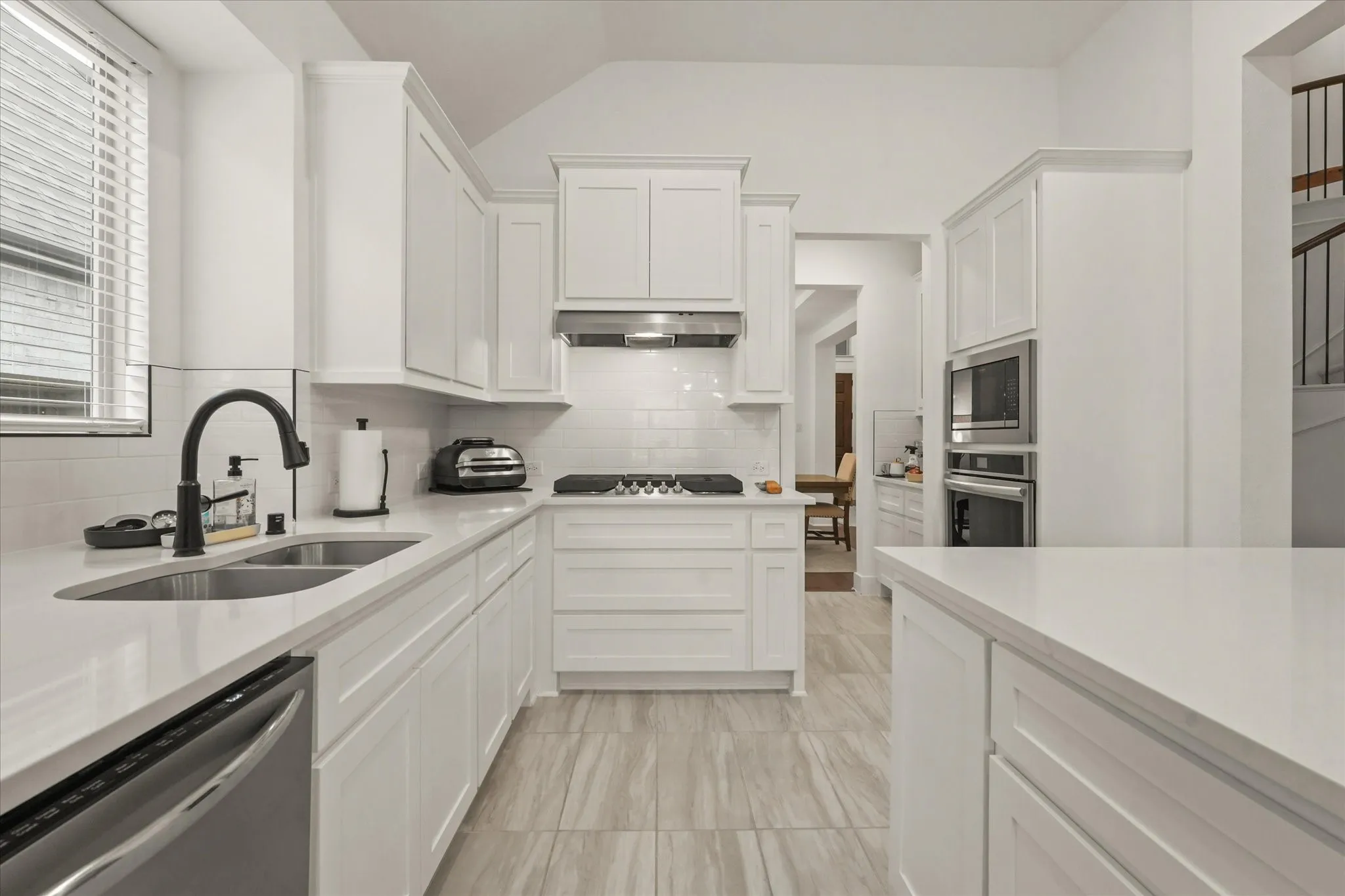 White shaker-style cabinetry with crown molding, stainless steel appliances, a black gooseneck faucet over a double basin sink, light-colored countertops, and wood-finish tile flooring