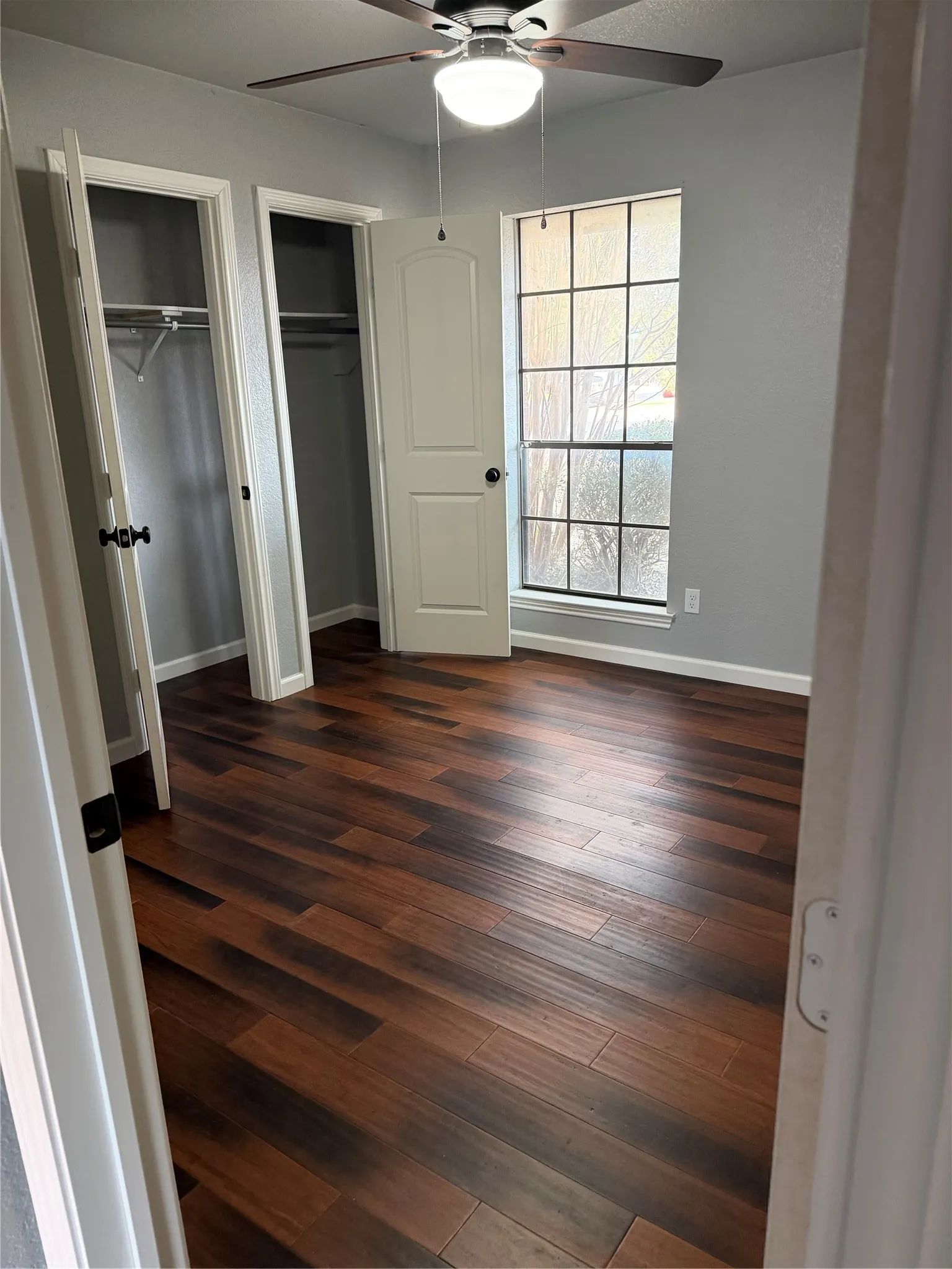 Wood-finish flooring, overhead ceiling fan with light fixture, window with grid pattern, and two built-in closets