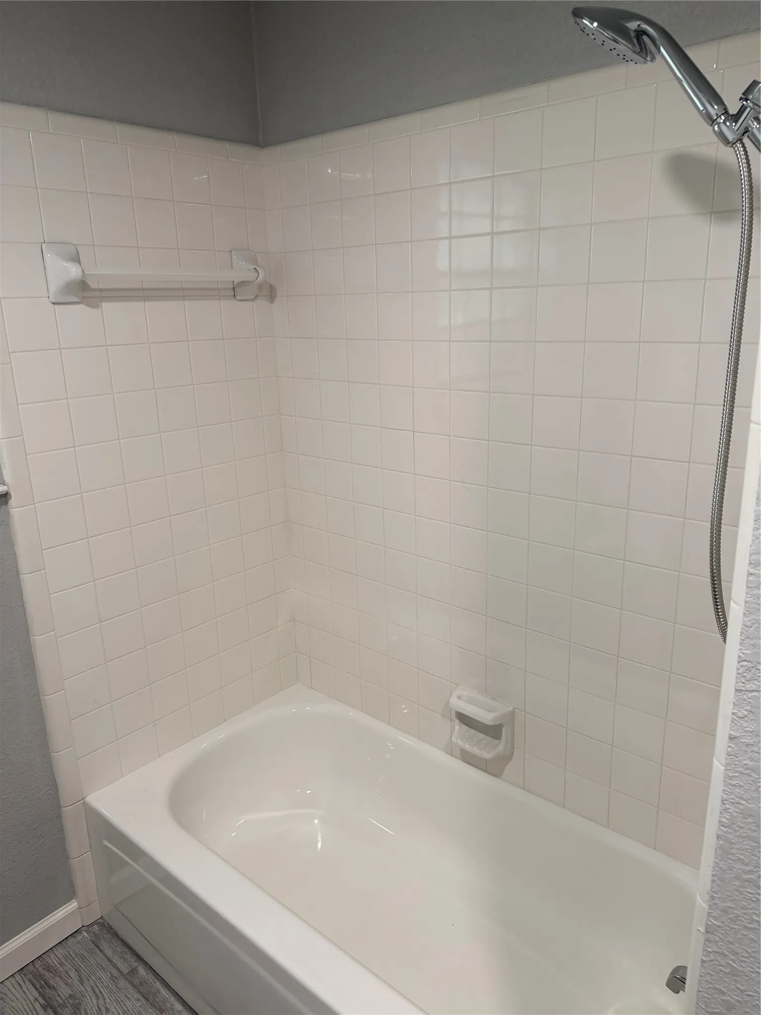Bathtub with white subway tile surround, chrome handheld shower fixture, and gray wood-finish flooring