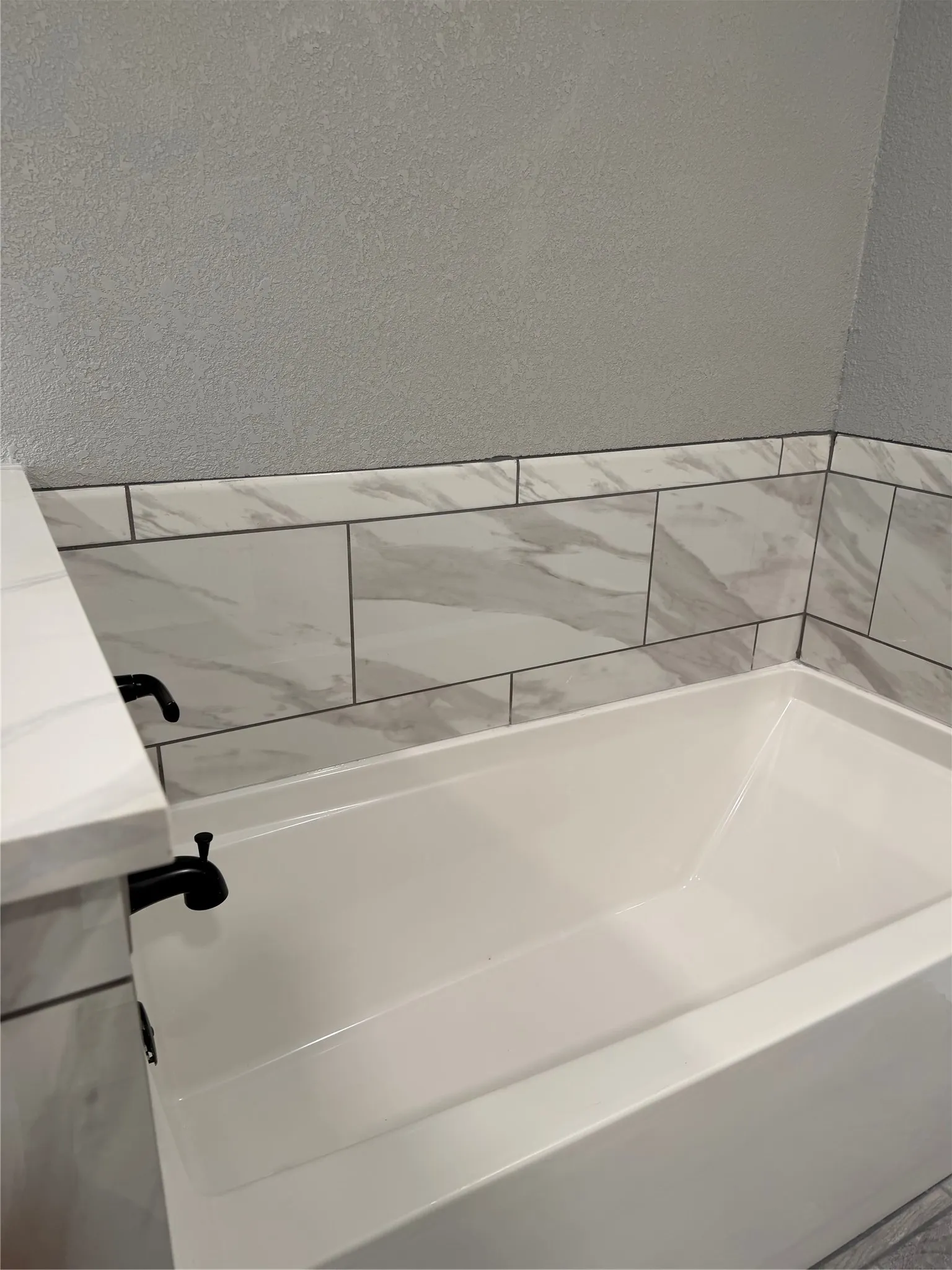 Sleek white tub with matte black fixtures, complemented by white and gray veined wall tiling and a coordinating countertop