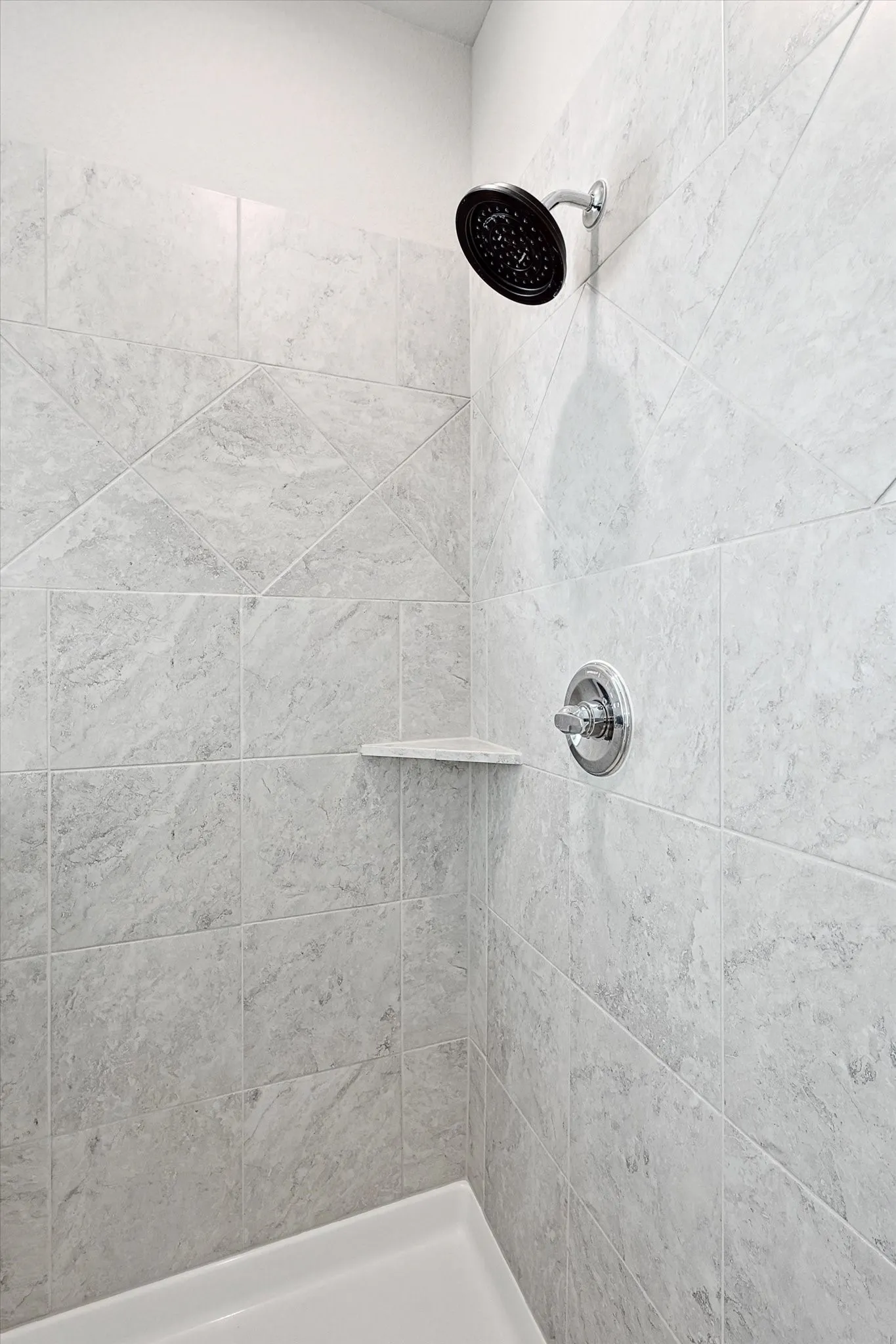 Shower enclosure featuring large format tile with geometric accents, a built-in corner shelf, dark bronze shower head, and chrome fixtures