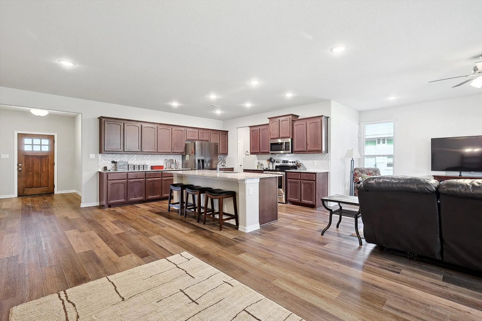 Open-concept living space with wood-finish flooring, a kitchen island with seating, dark wood cabinetry, stainless steel appliances, and recessed lighting