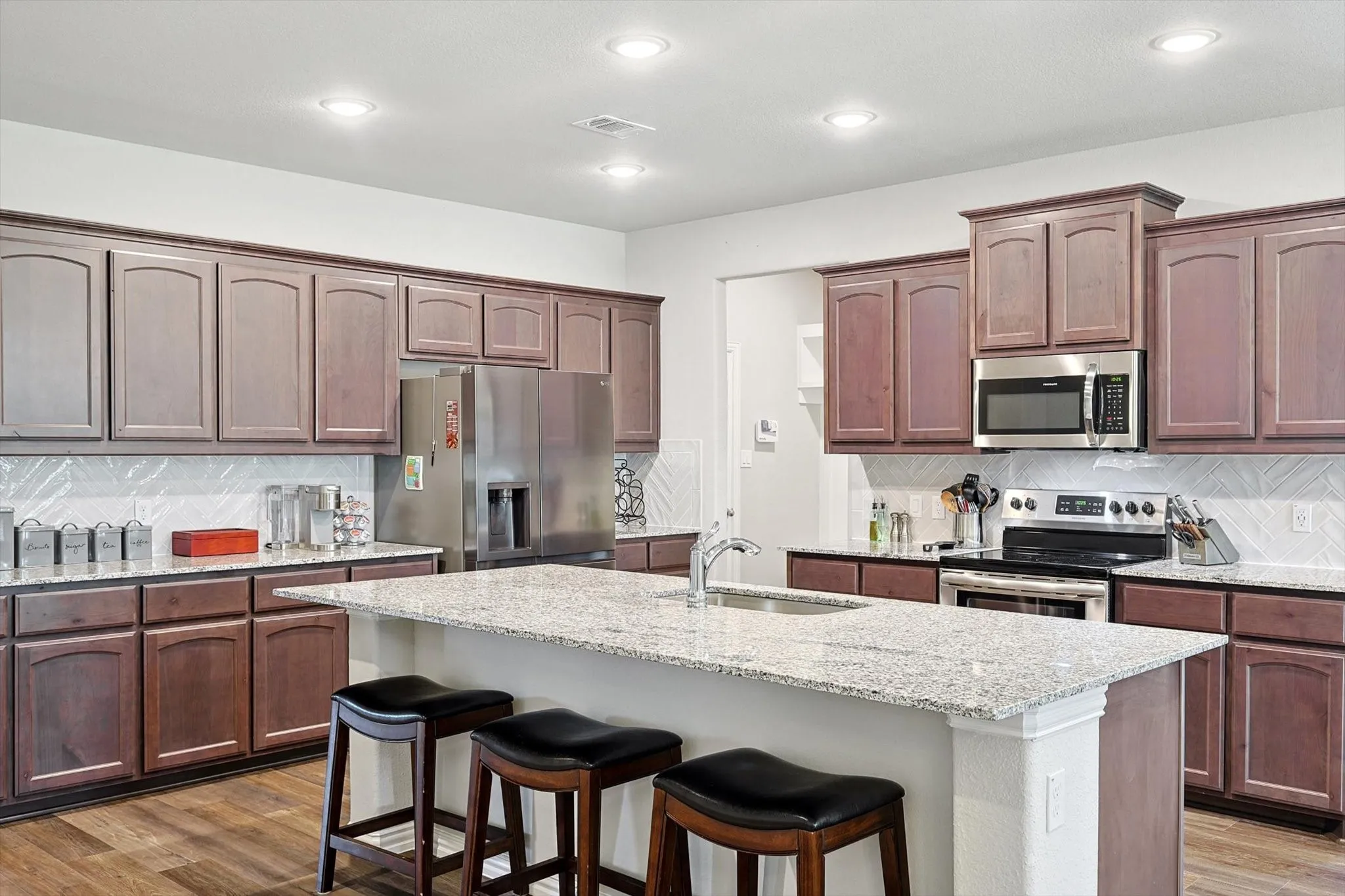 Kitchen featuring a center island with sink, shaker-style cabinetry, stainless steel appliances, granite countertops, and wood-finish flooring