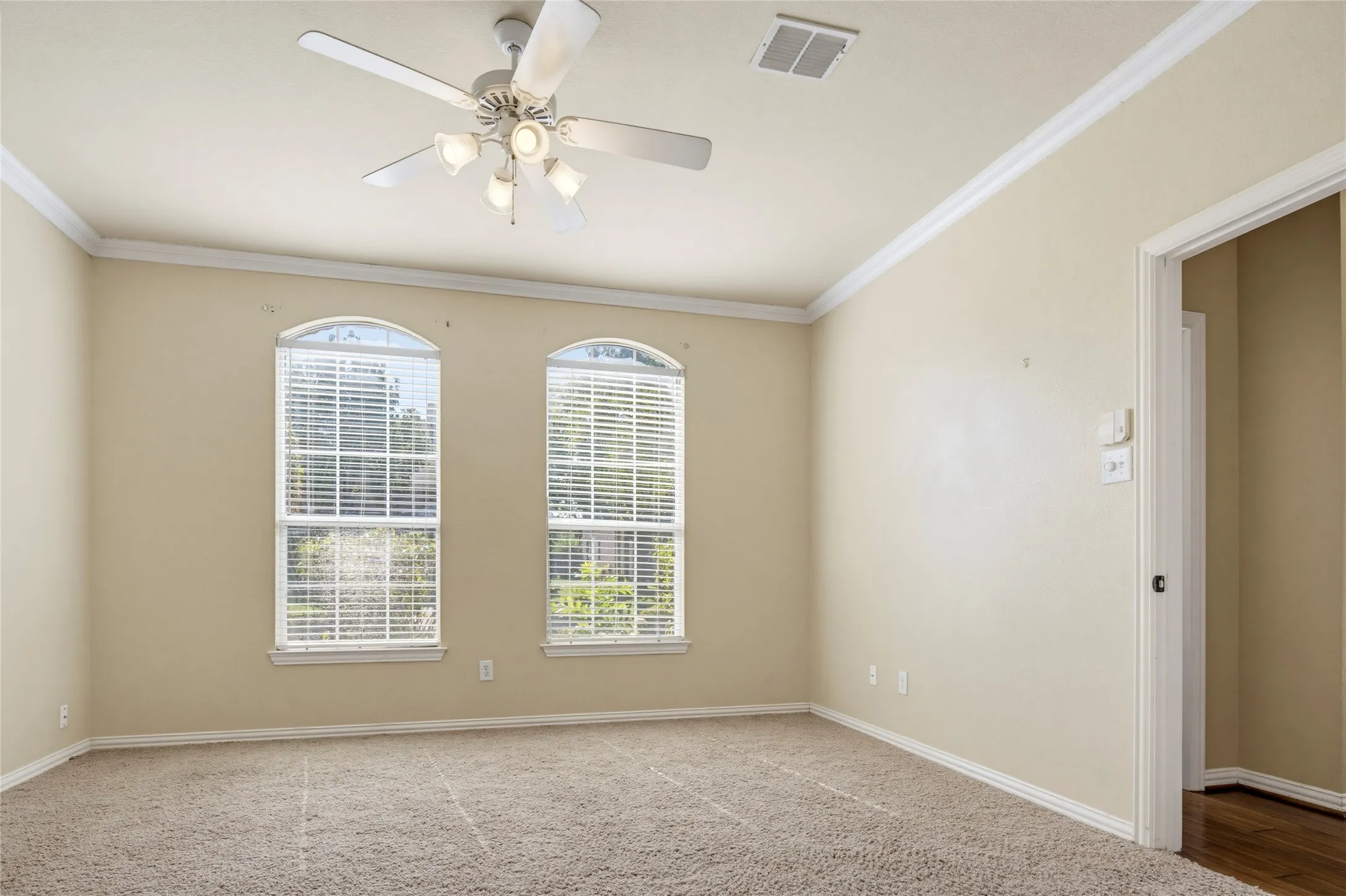 Carpeted flooring complemented by crown molding, arched windows with blinds, a ceiling fan, and a doorway with wood-finish flooring beyond