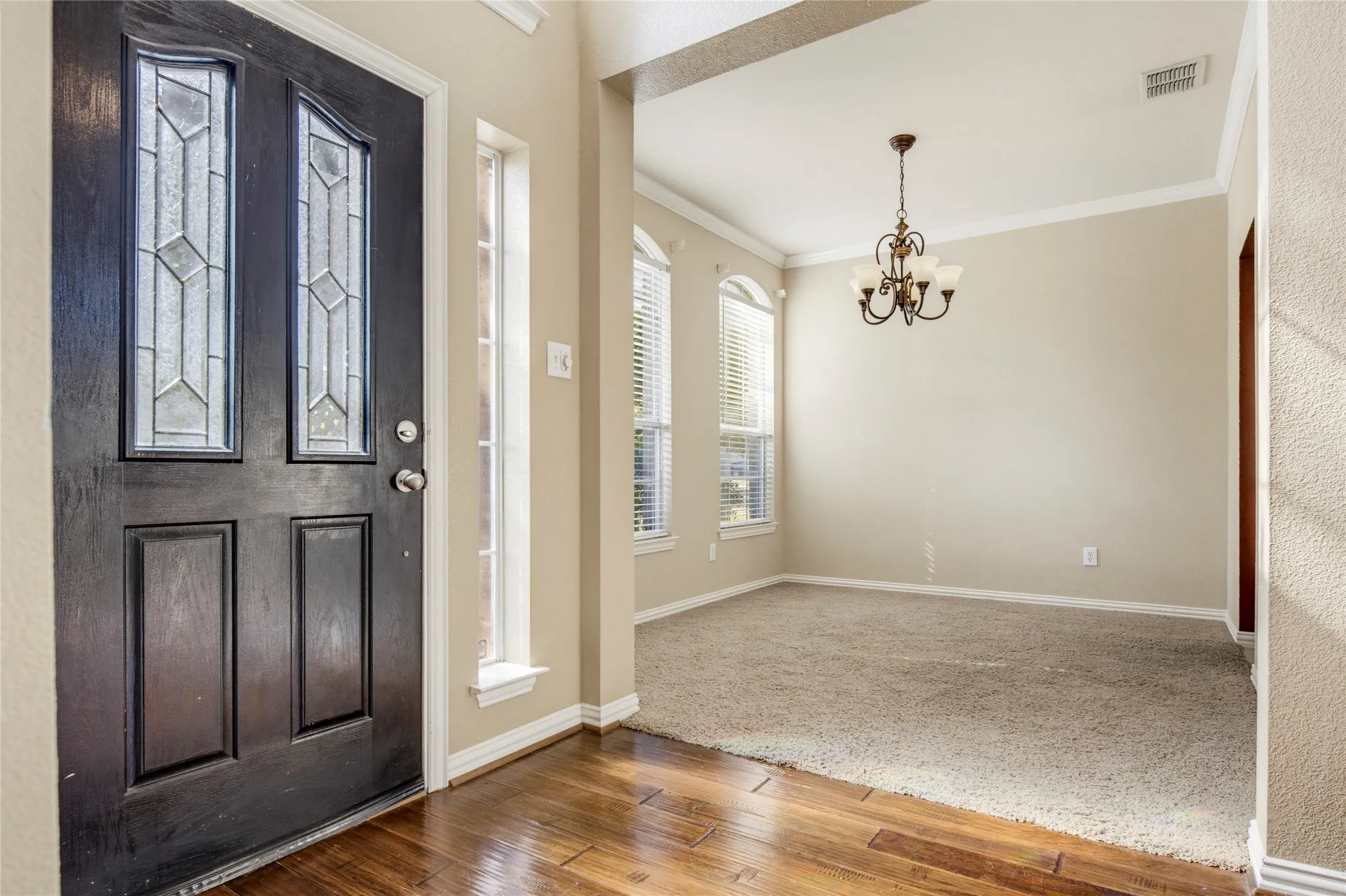 Dark wood entry door with decorative glass inserts, wood-finish flooring, and a chandelier in the adjacent room with arched windows and carpet