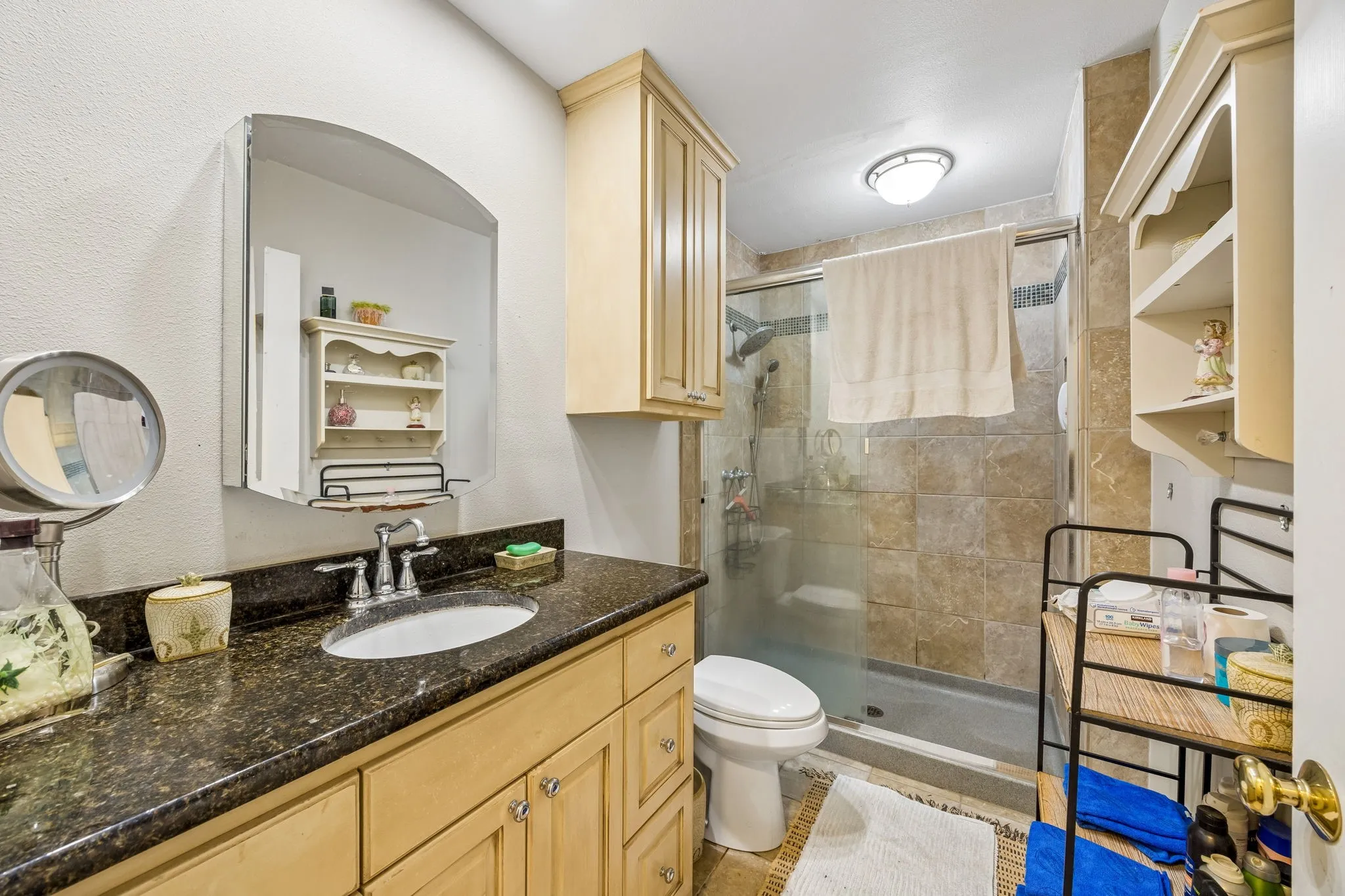 Bathroom featuring a vanity with a dark speckled countertop, light wood-finish cabinetry, a framed mirror, a tiled shower with a glass enclosure, and a built-in shelving unit