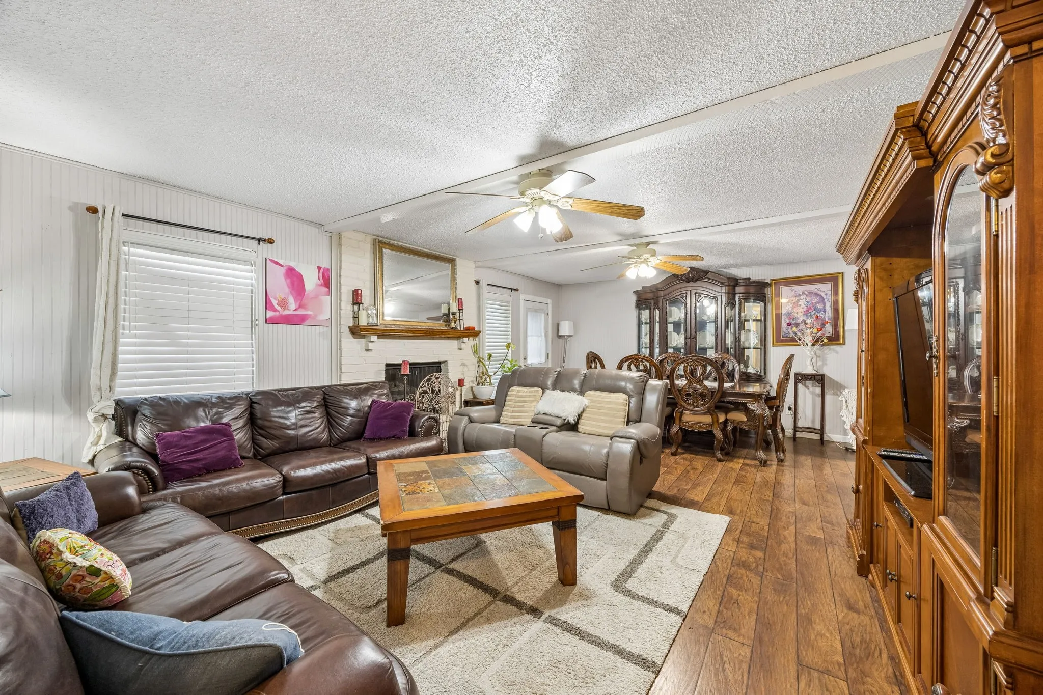 Spacious living area featuring wood-finish flooring, a white brick fireplace with a mantel, and built-in ceiling fans