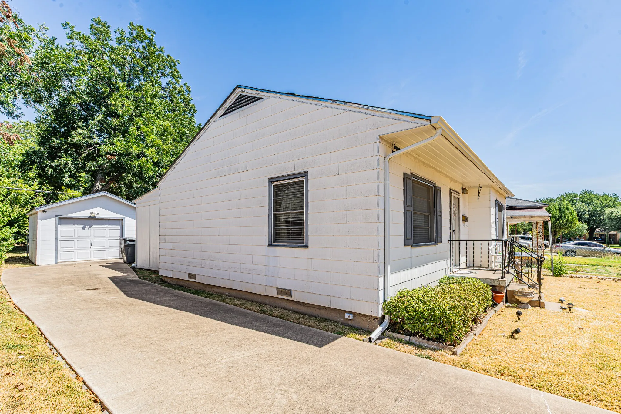 White siding exterior with a pitched roof, concrete driveway, detached garage with roll-up door, and covered front porch with iron railing