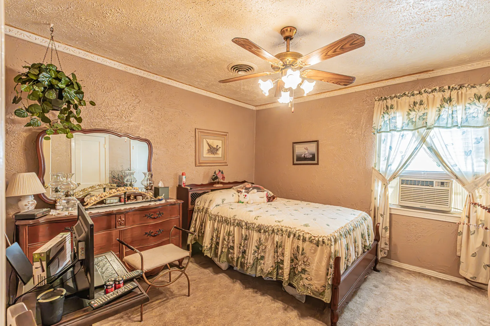 Textured wall finish, ceiling fan with integrated lighting, window-mounted air conditioning unit, window with valance and curtains, and wood-finish bed frame