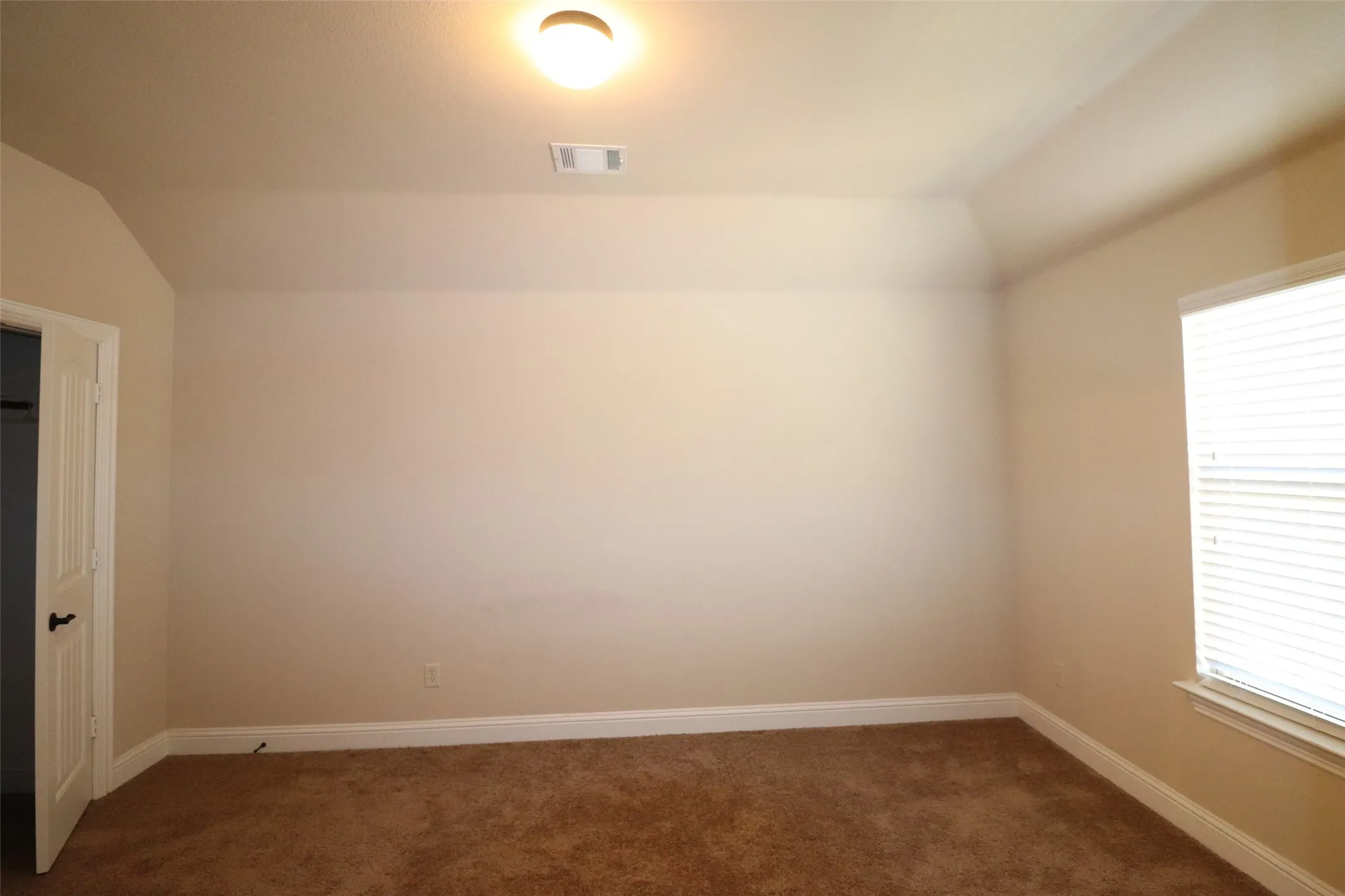 Vaulted ceiling, wall-to-wall carpeting, window with blinds, interior door with dark hardware, flush-mount ceiling light