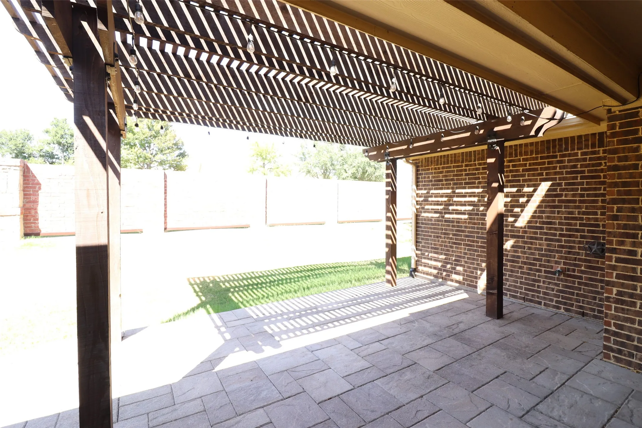 Expansive outdoor patio featuring a wooden pergola, brick exterior wall, and patterned stone-look paving