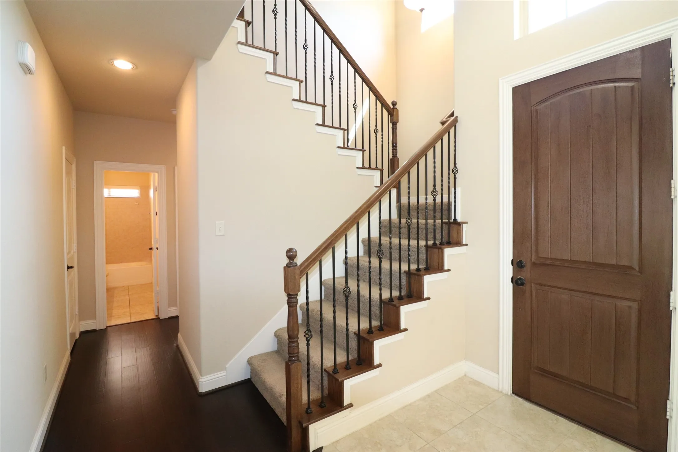 Wood-finish entry door, tile flooring, wood-finish staircase with wrought iron balusters, recessed lighting, and dark wood-finish flooring in the hallway