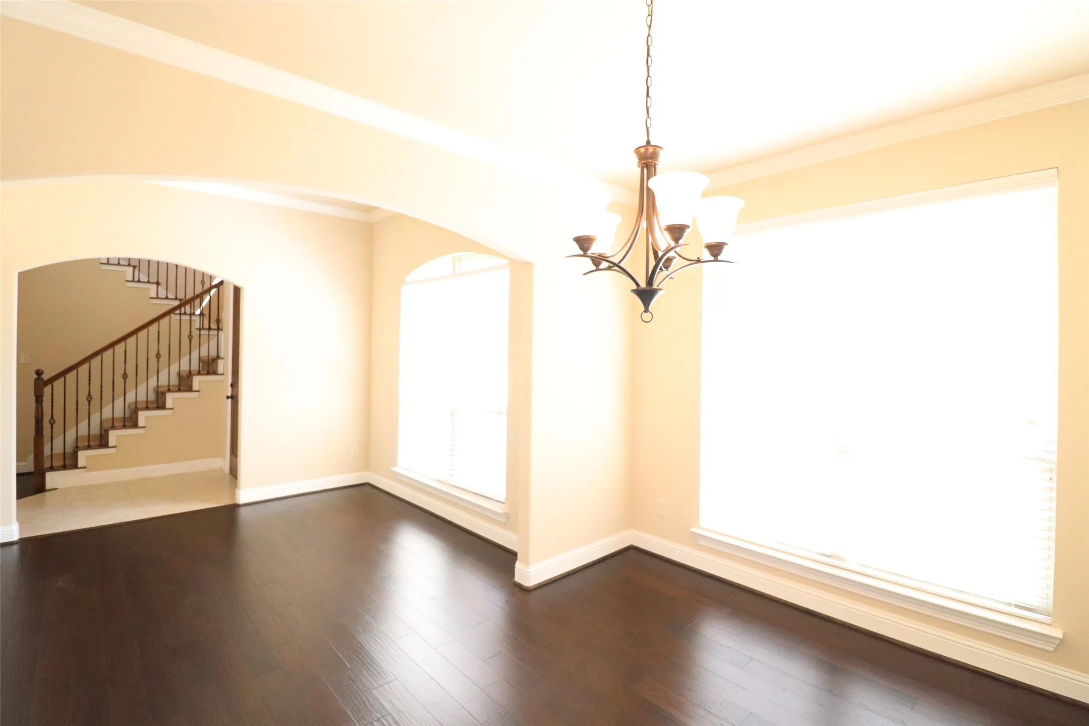 Dark wood-finish flooring, two large windows with blinds, arched entryways, and a staircase with an iron railing